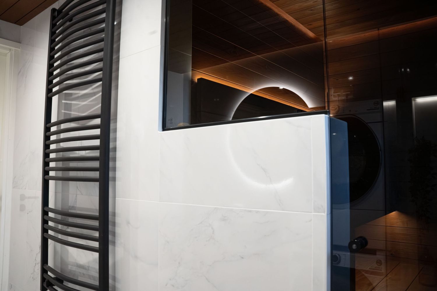 Modern bathroom with marble tiles, black heated towel rail, wooden ceiling, and round backlit mirror.