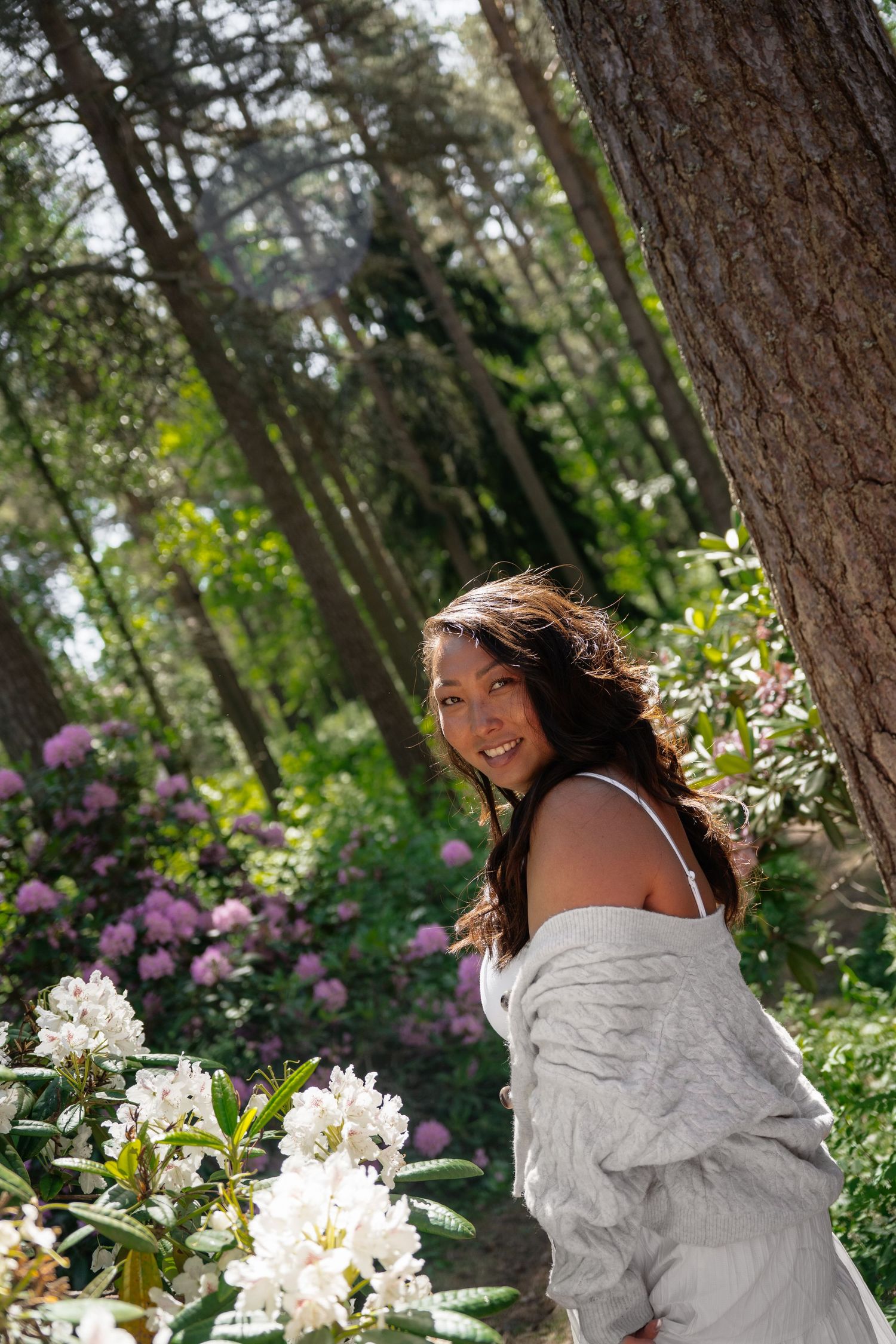 Smiling woman in white dress posing among blooming rhododendrons in a sunlit forest.
