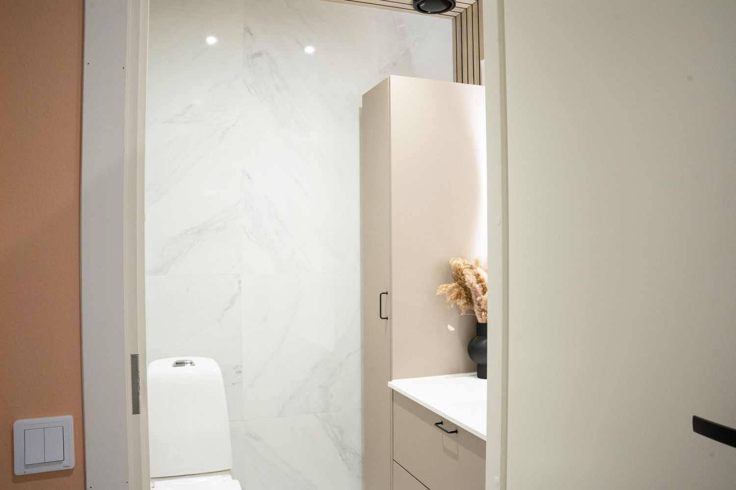 Modern bathroom with white marble walls, tall storage cabinet, and toilet visible through open door.