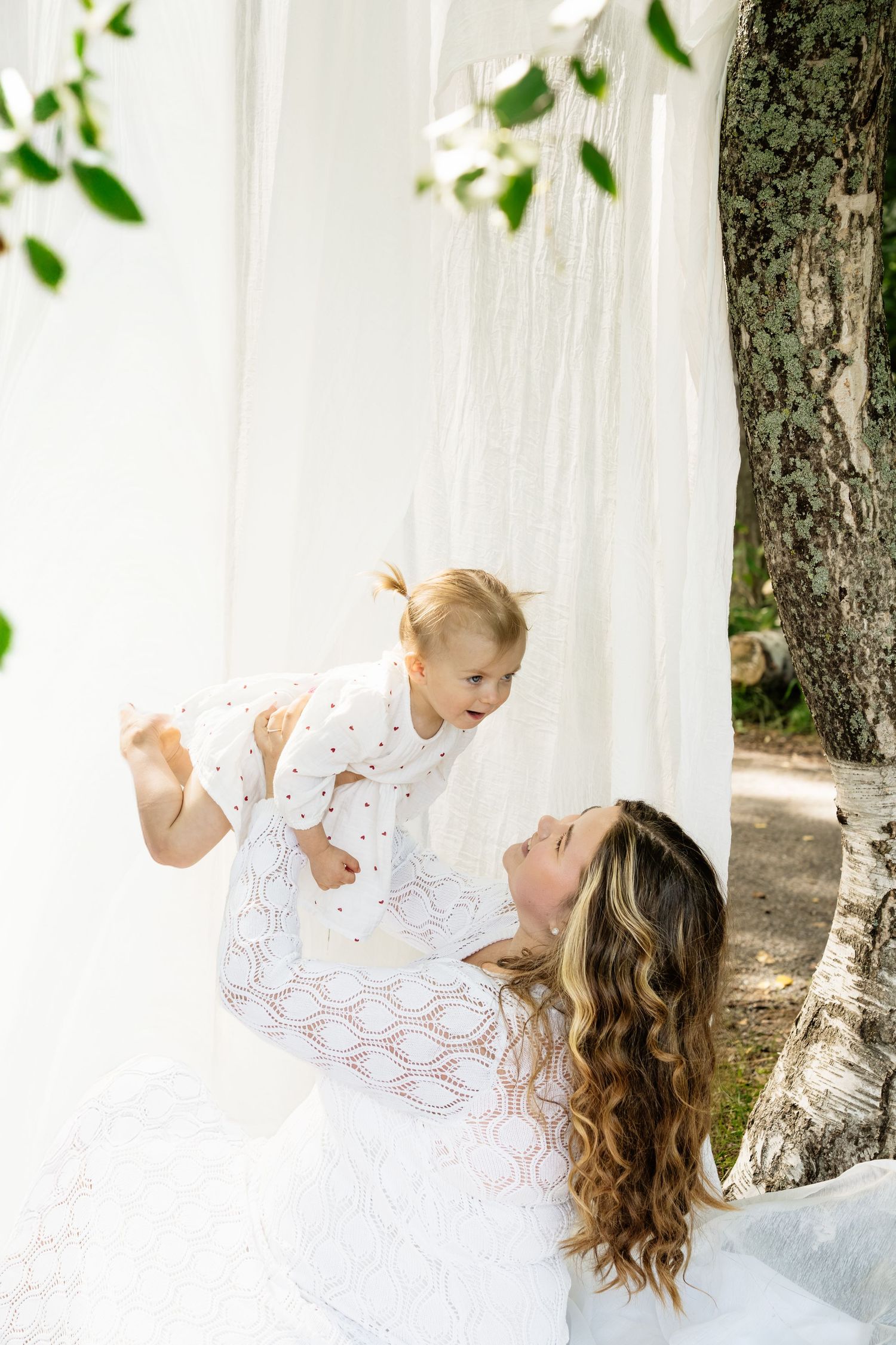 A mother in a white lace dress joyfully lifts her laughing toddler outdoors near white curtains and a tree.