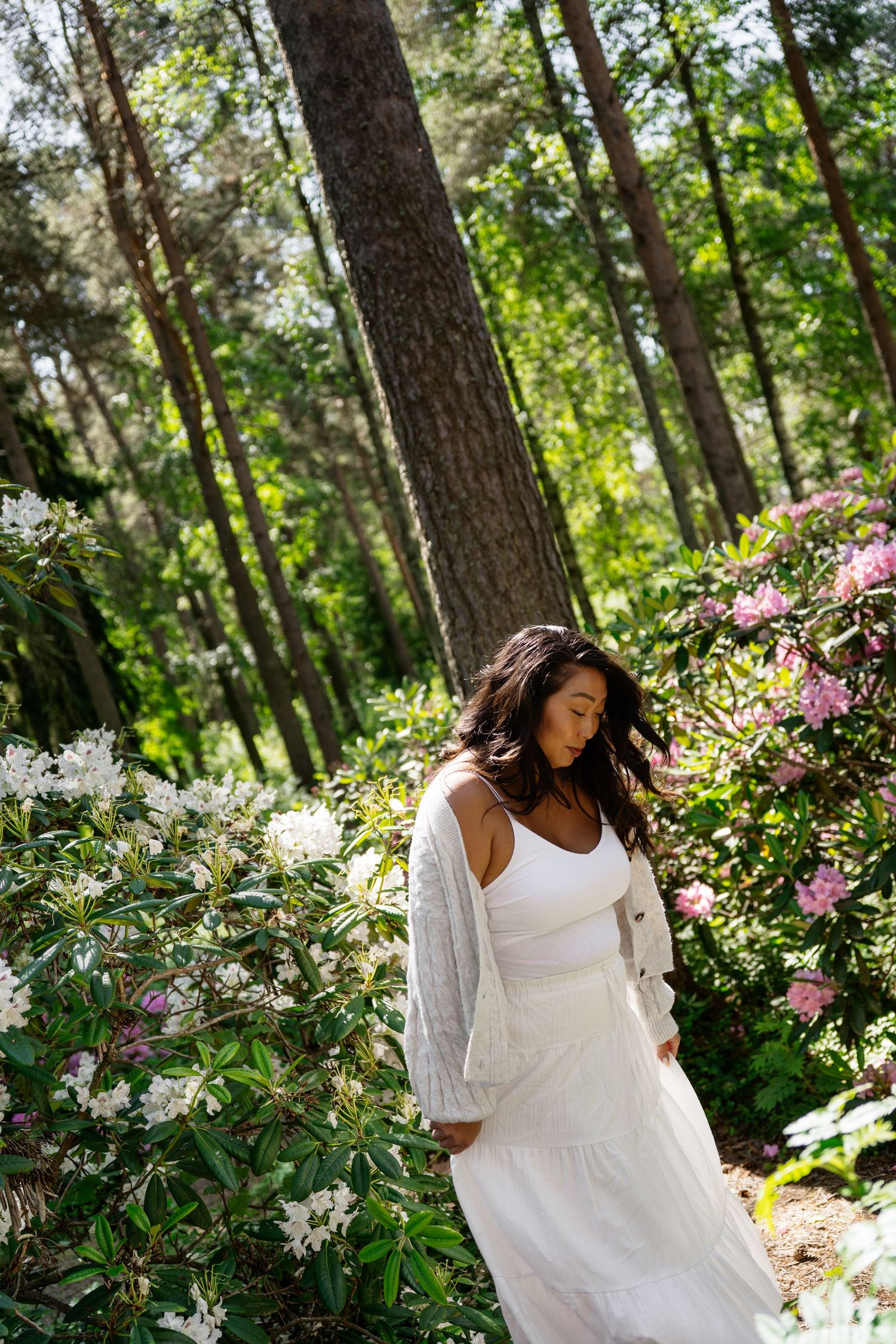 Woman in white dress and cardigan standing among blooming flowers in a sunlit forest setting.