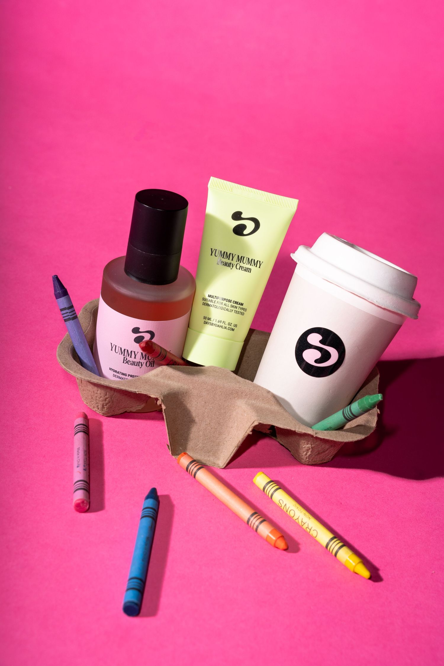 Yummy Mummy beauty products displayed with colorful crayons in a cardboard tray on a vibrant pink background.
