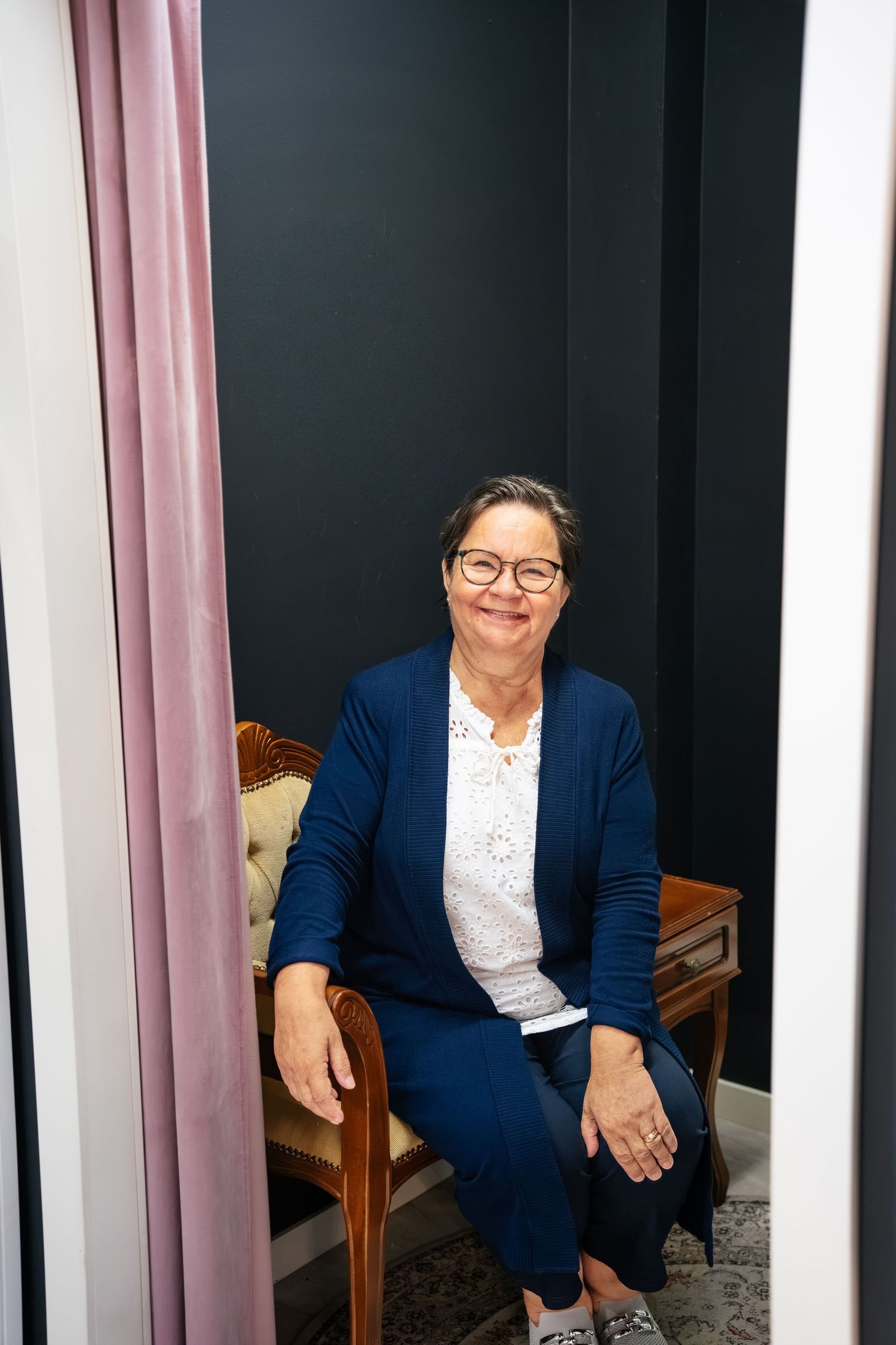 Woman in blue blazer smiling while seated in a fitting room with pink curtain and dark walls.