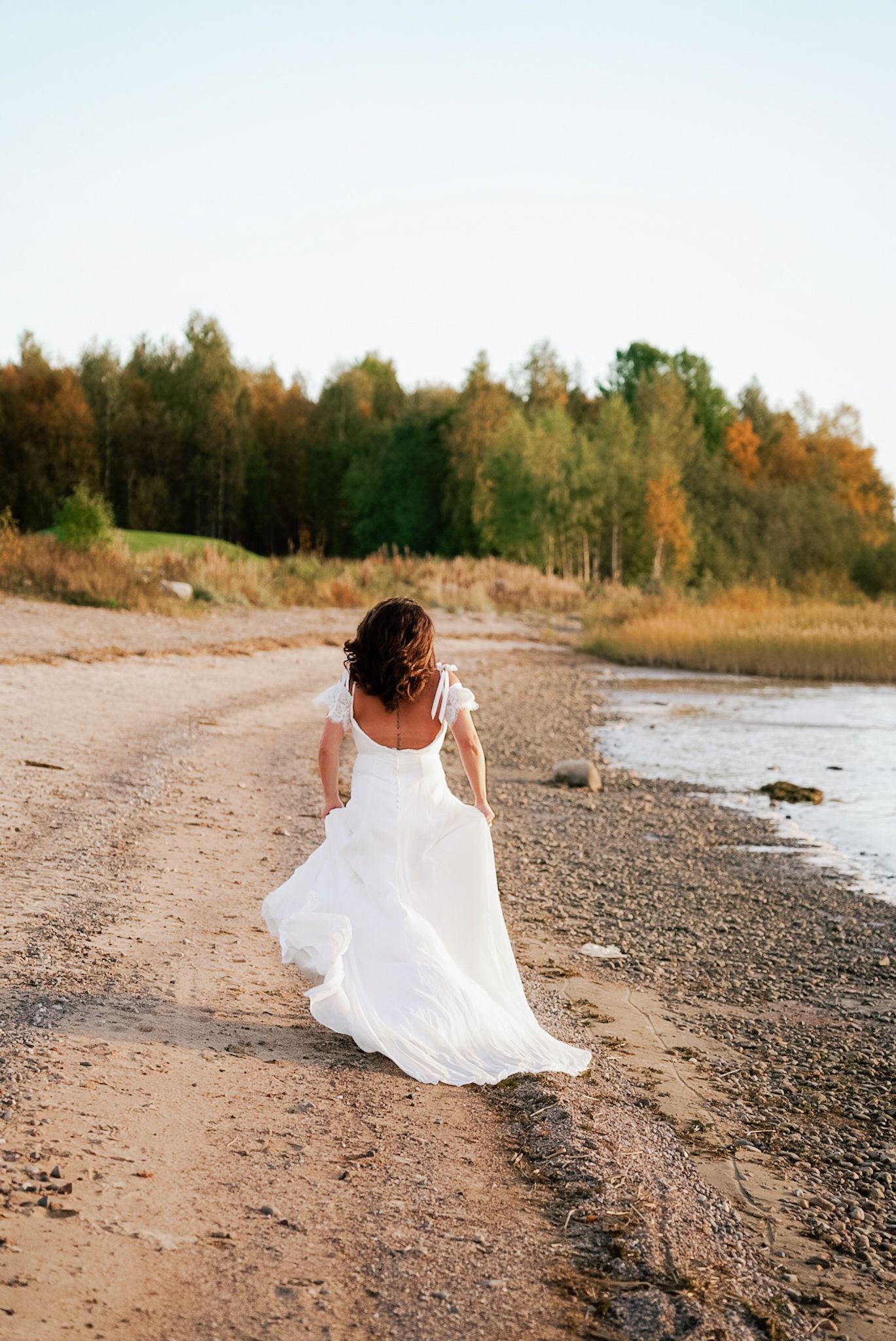 Bride in a flowing white wedding dress walking along a sandy beach with autumn trees in background.