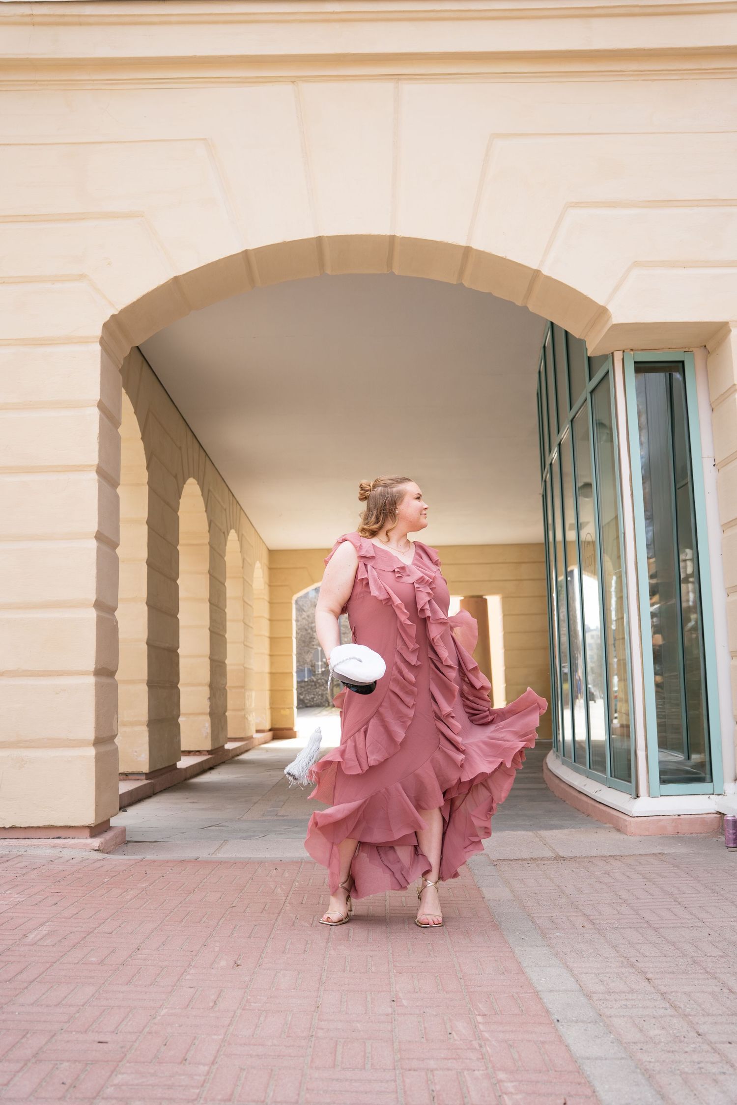 Woman in flowing pink ruffle dress walks barefoot under stone arch, holding white hat, at elegant building exterior.