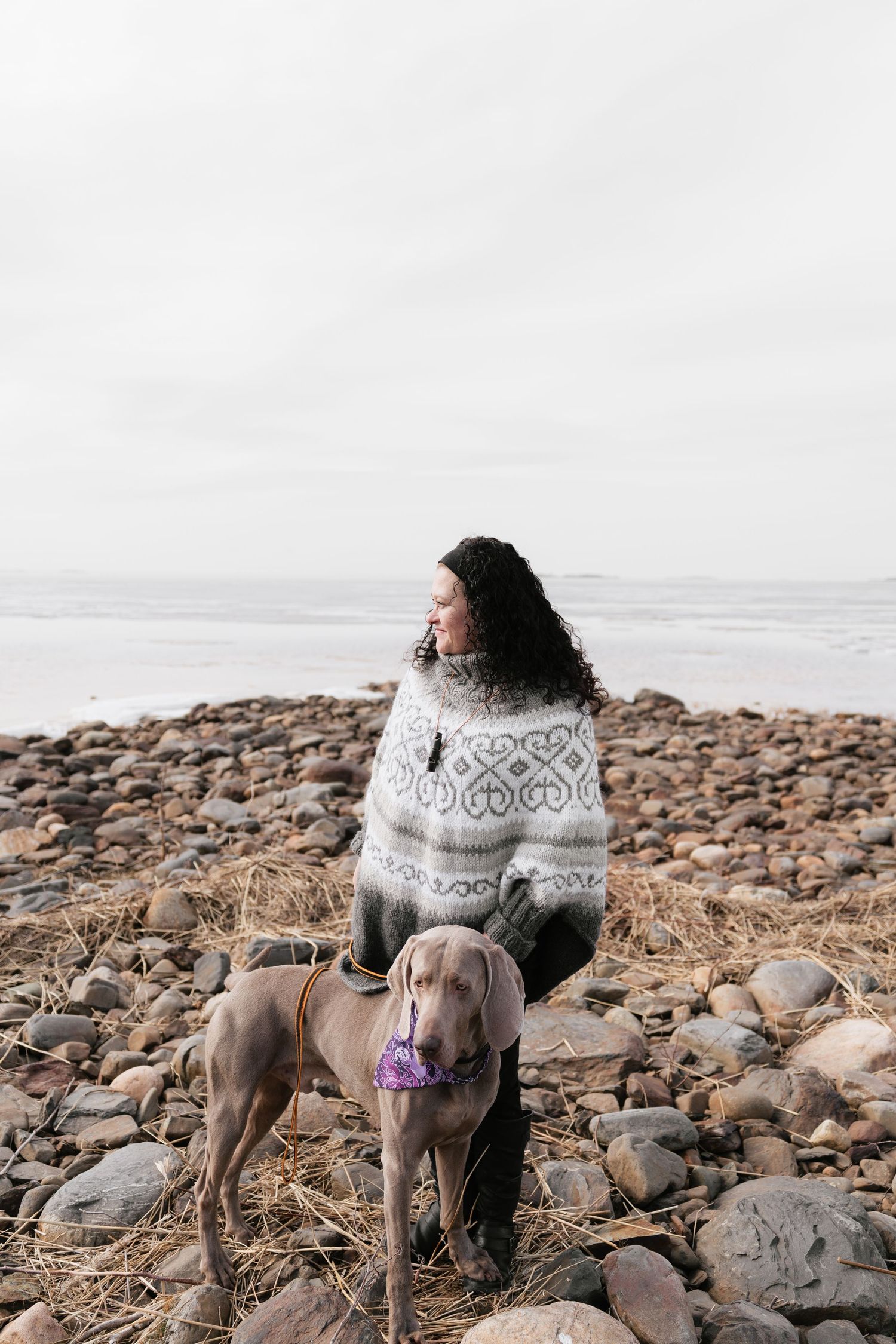 Woman in Nordic sweater sits on rocky beach with Weimaraner dog wearing purple bandana, overcast sky.