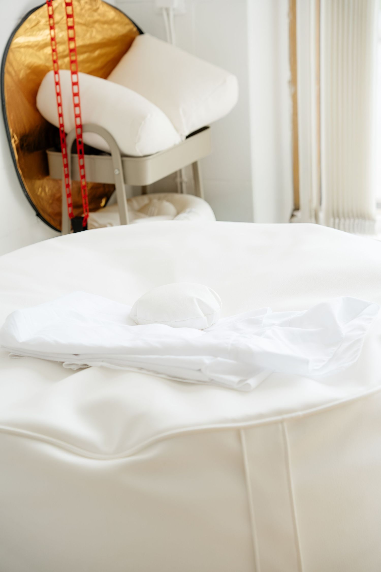 White folded bed linens on a table with a chair and red beaded necklace reflected in a round mirror.
