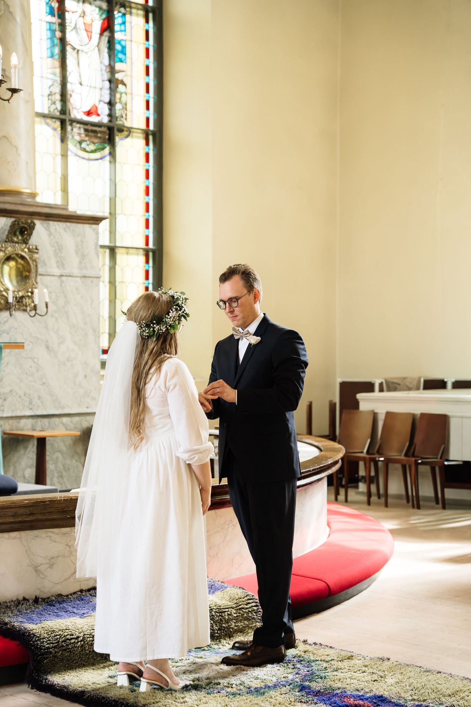 Bride and groom exchange rings at church altar, bride wearing floral crown and white gown, groom in dark suit.