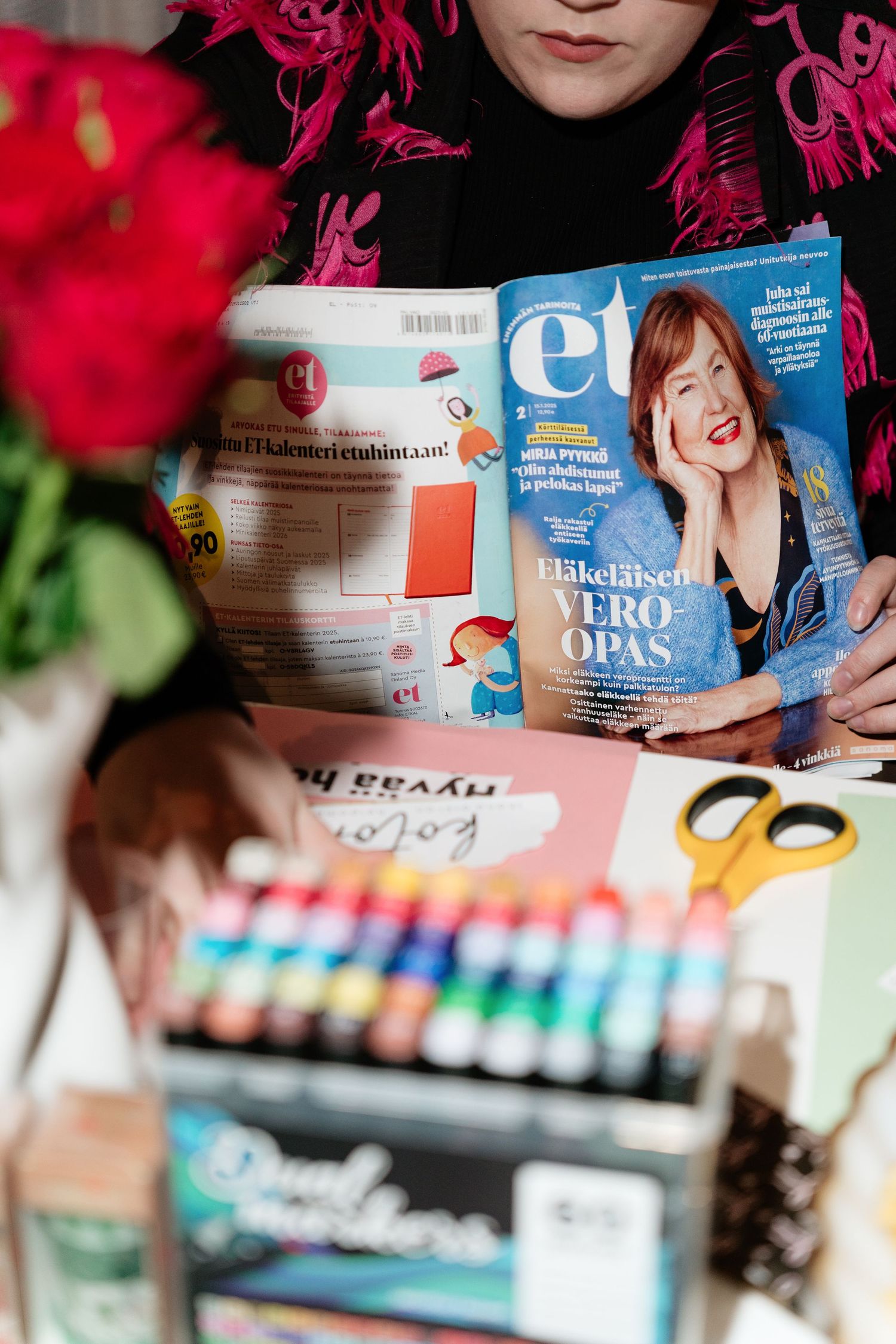 Person holding ET magazine with Eläkeläisen Vero-opas cover, art supplies and red flowers in foreground.