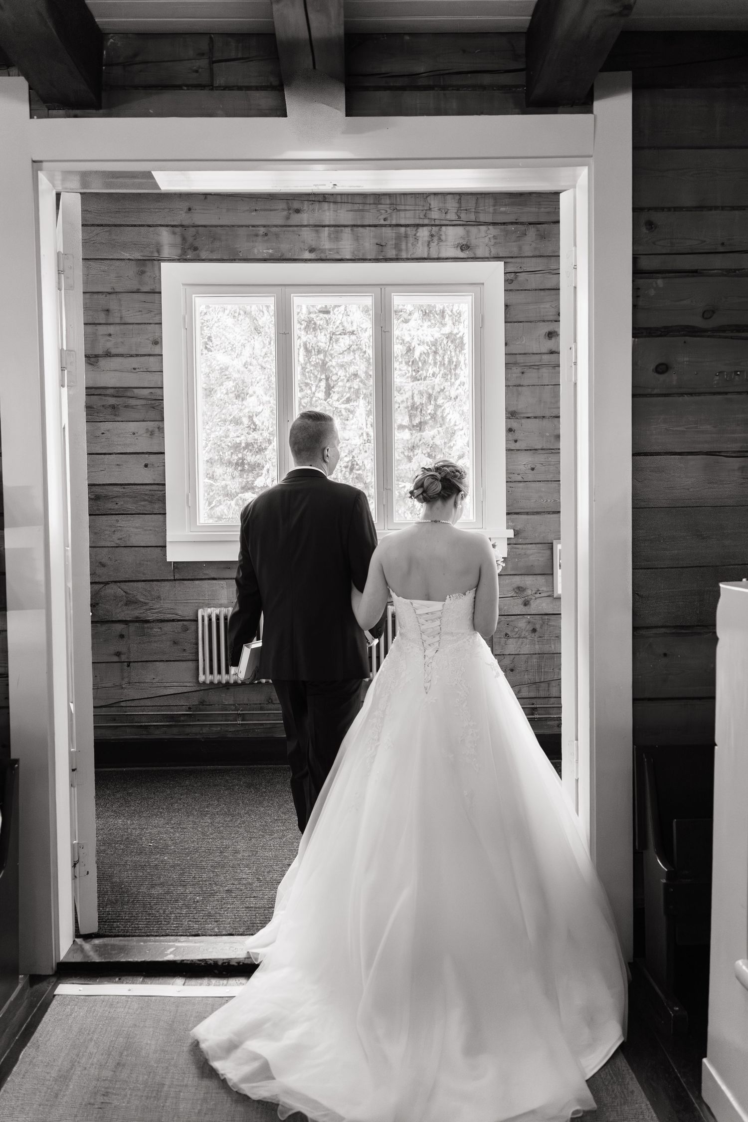 Black and white photo of bride and groom holding hands, walking toward a window in a rustic wood-paneled room.