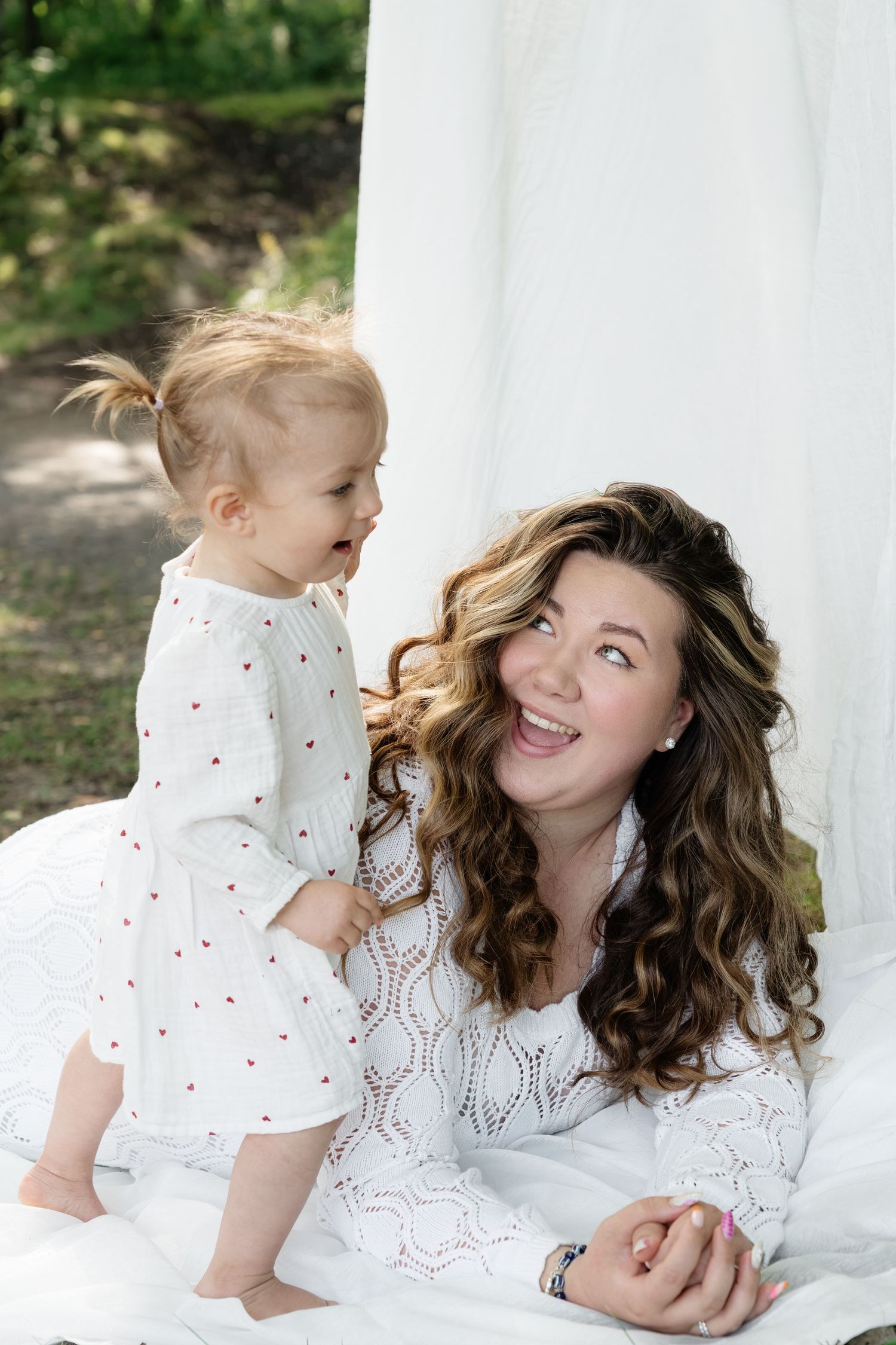 Laughing mother in white lace dress sits with standing toddler girl in white heart-print dress outdoors.
