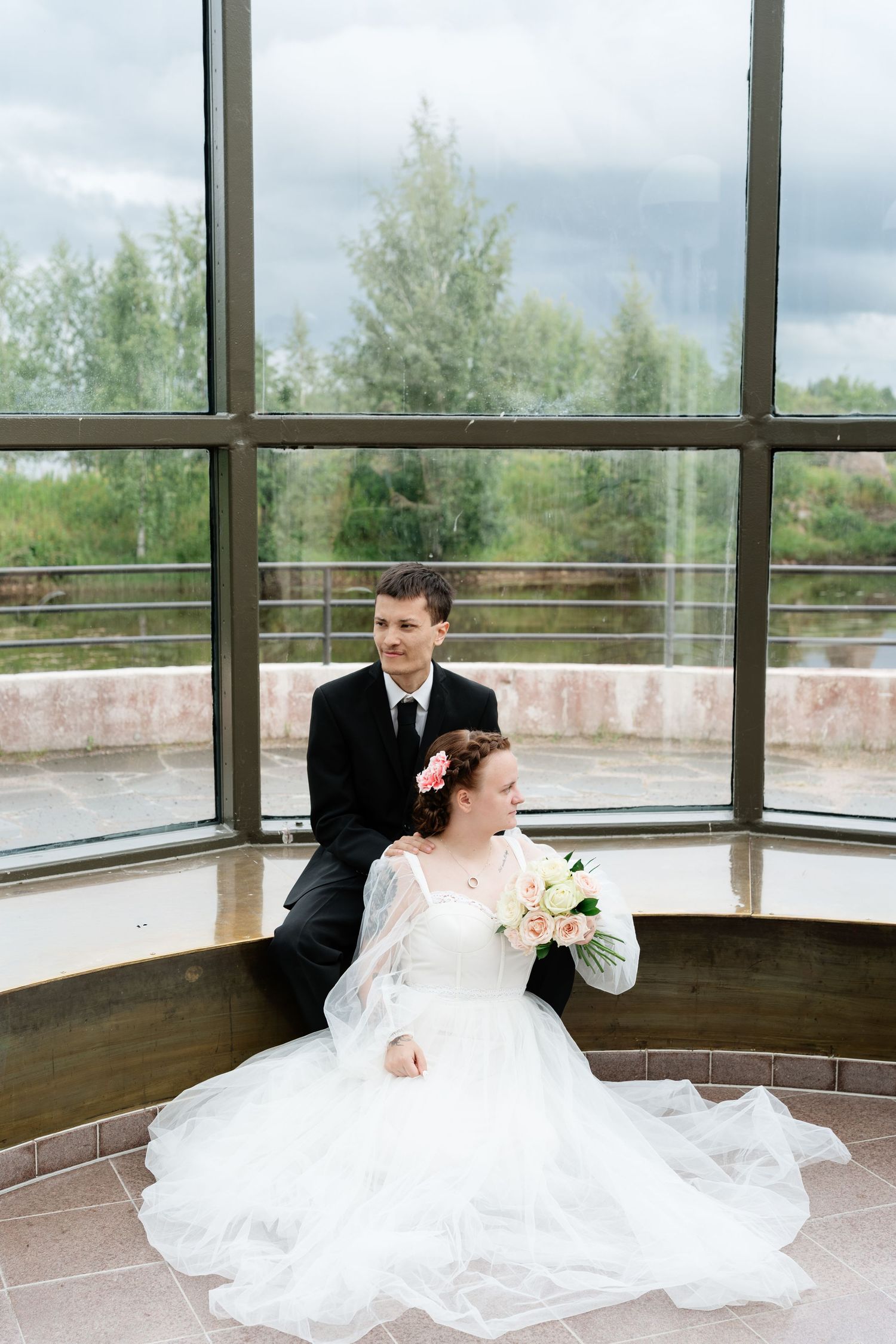 Bride in white gown and groom in black suit sitting by large window on their wedding day.