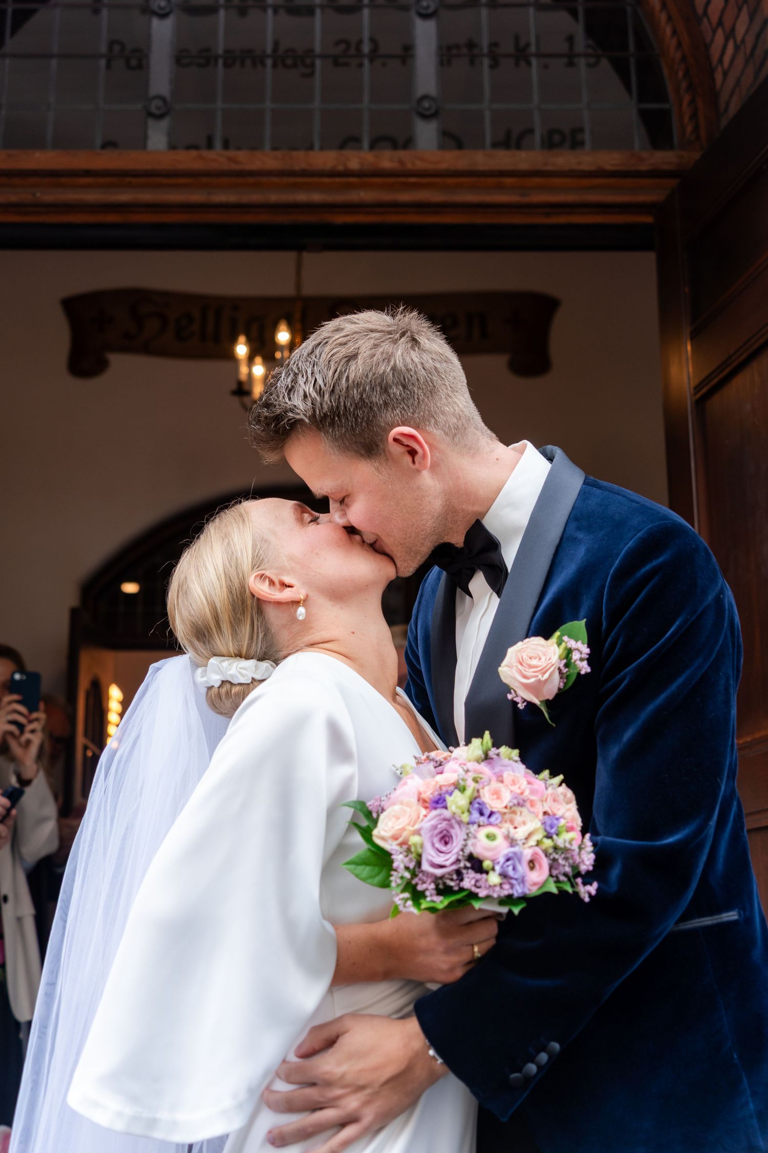 Bride and groom share a kiss outside a church, bride holding a pink and purple bouquet, groom in a navy blue velvet suit.