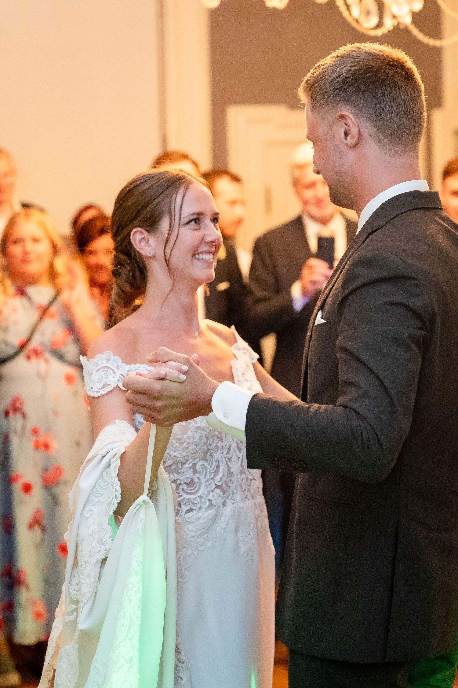 Bride and groom share their first dance, smiling joyfully, surrounded by wedding guests in elegant attire.