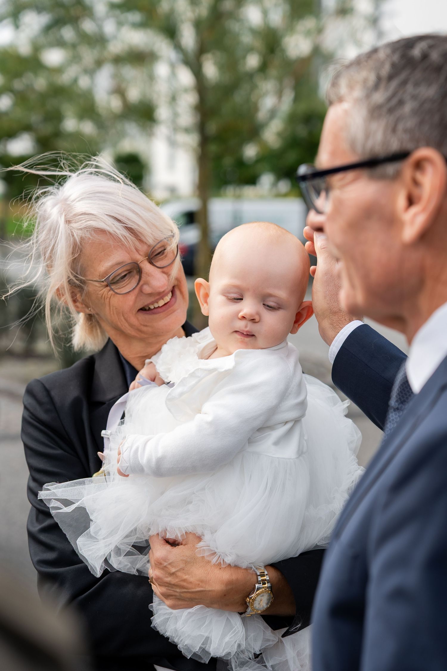 Grandparents smiling and holding a baby dressed in white at a christening ceremony outdoors.