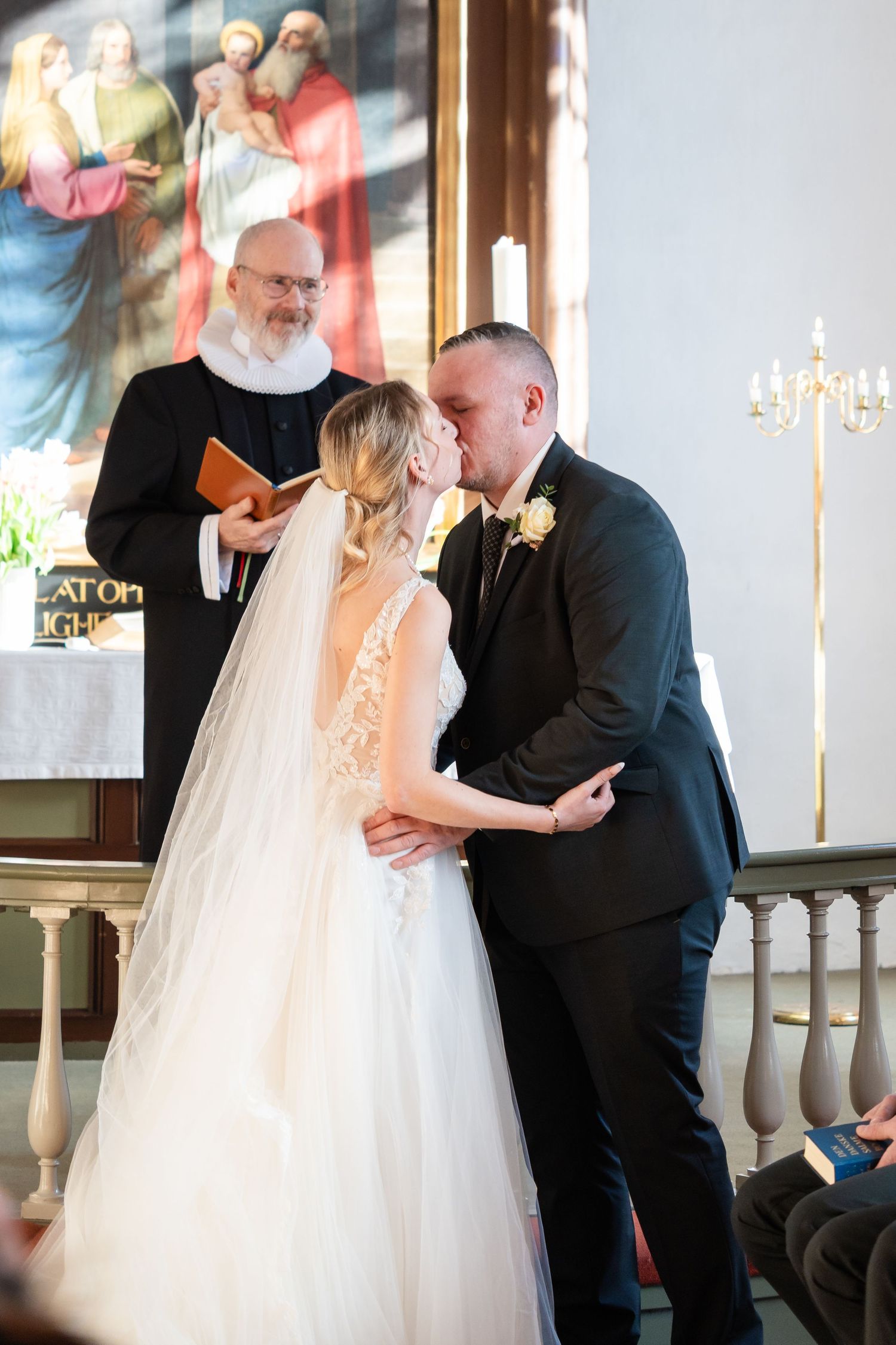 Bride and groom share first kiss at church altar during wedding ceremony with officiant holding book behind them.
