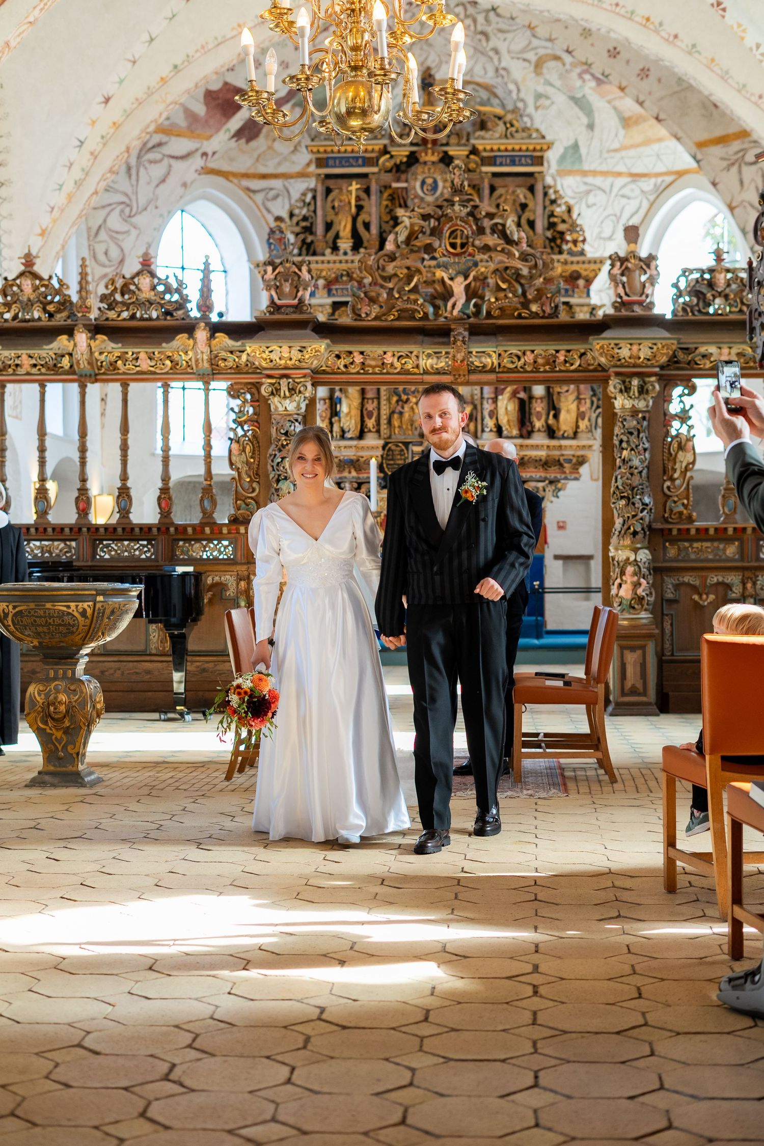 Bride and groom walking down the aisle of an ornate church with a golden baroque altar backdrop.