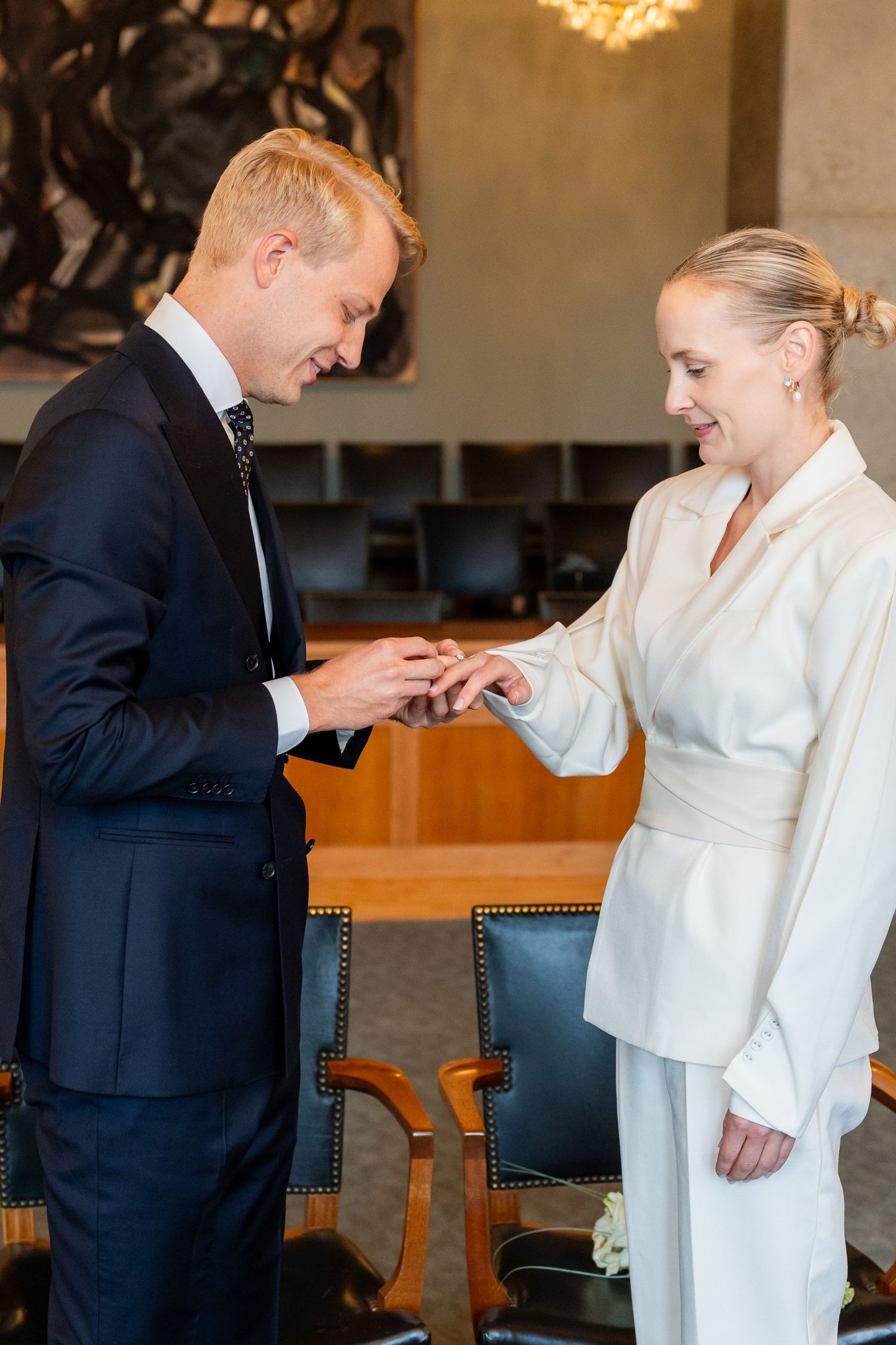 Groom in navy suit placing wedding ring on smiling bride in white outfit during intimate indoor ceremony.