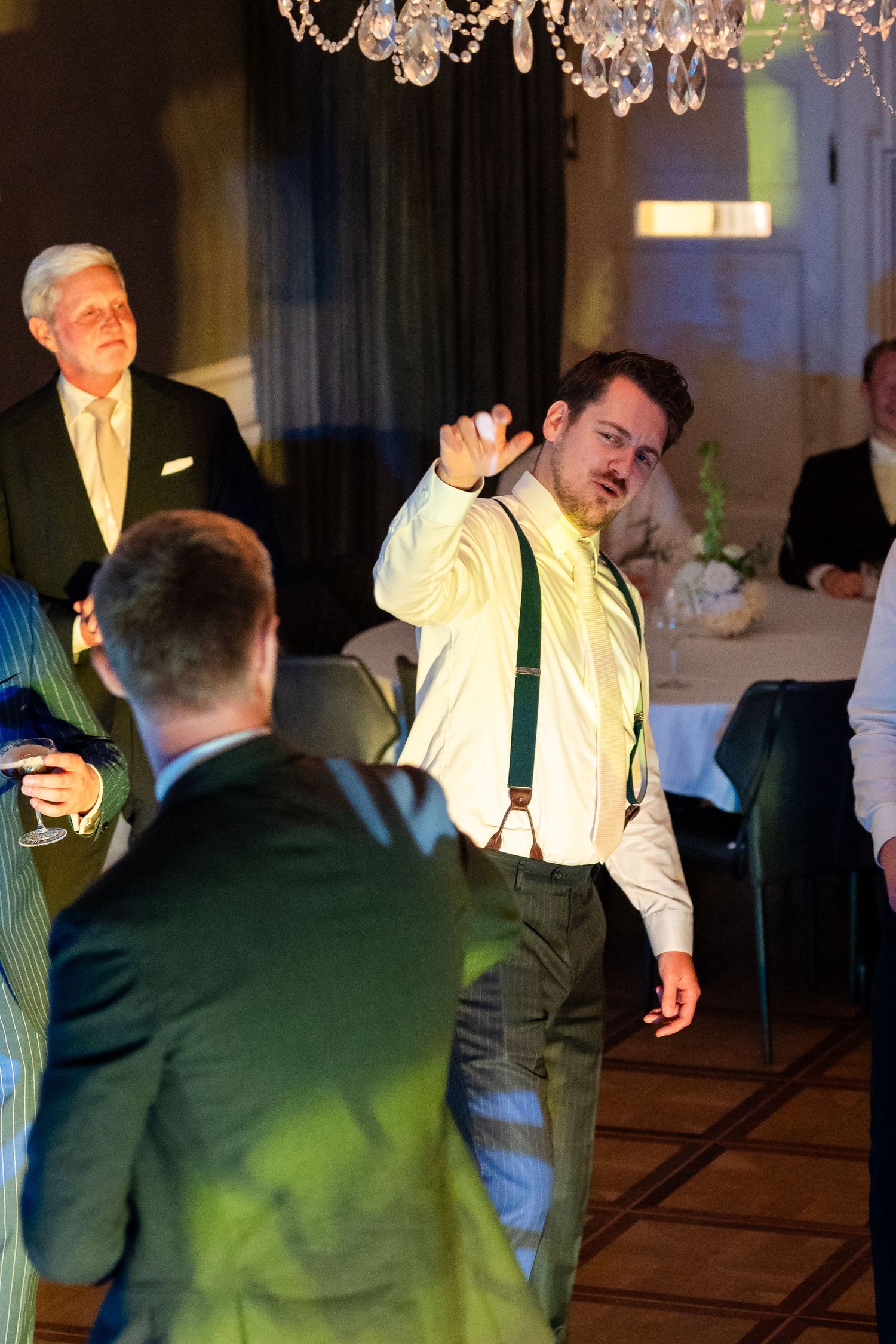 Man in yellow shirt and green suspenders dancing at an elegant indoor event with chandelier overhead.
