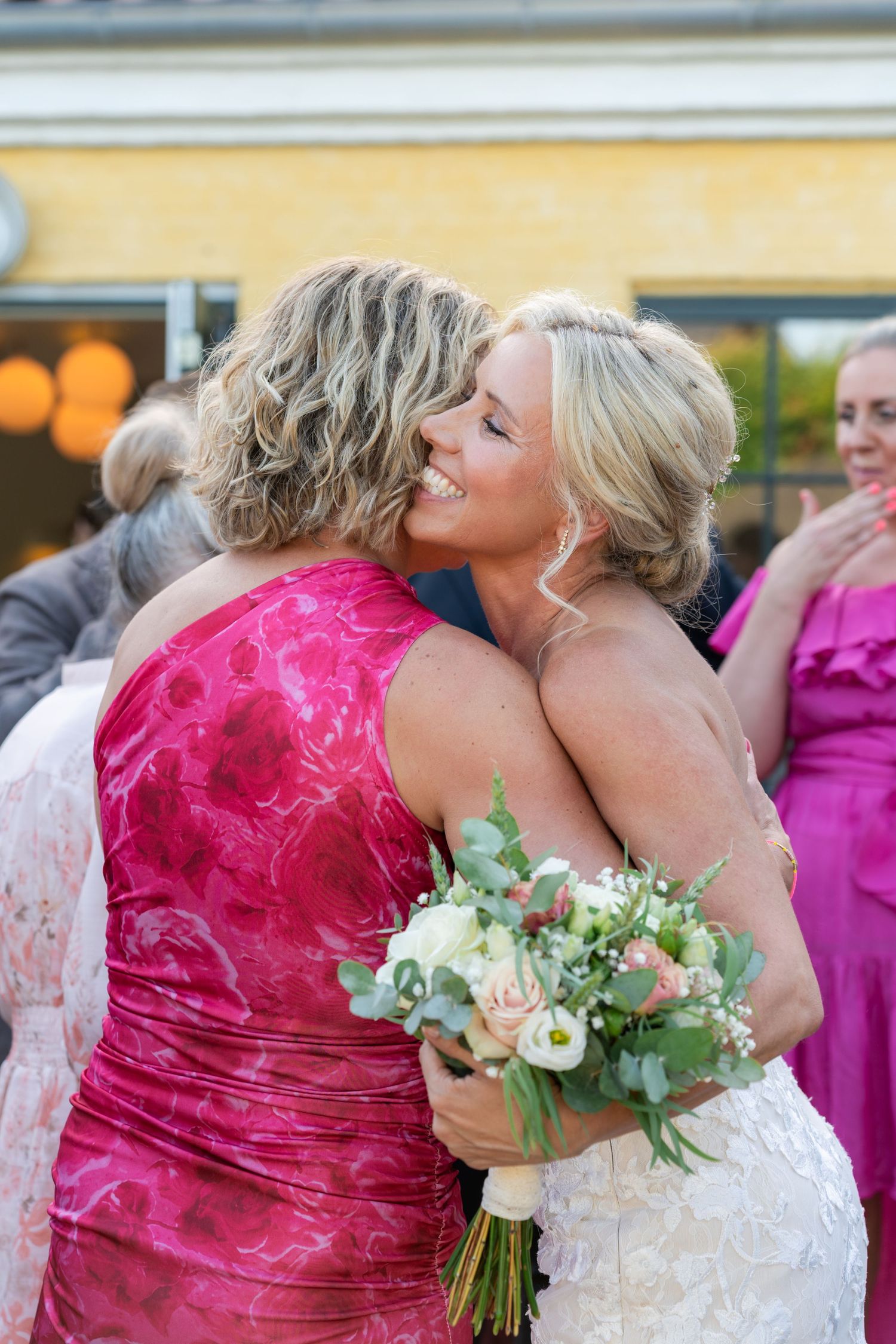Smiling bride in white gown hugging woman in pink floral dress while holding bouquet of roses and greenery at wedding.