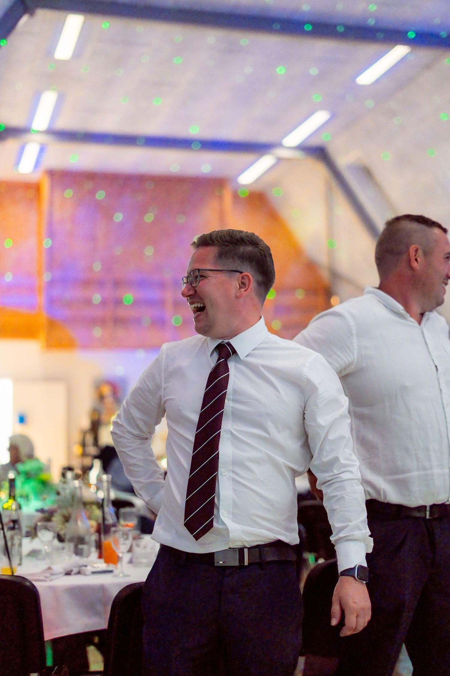 Man in white shirt and striped tie laughing at a formal event with colorful laser lights in background.