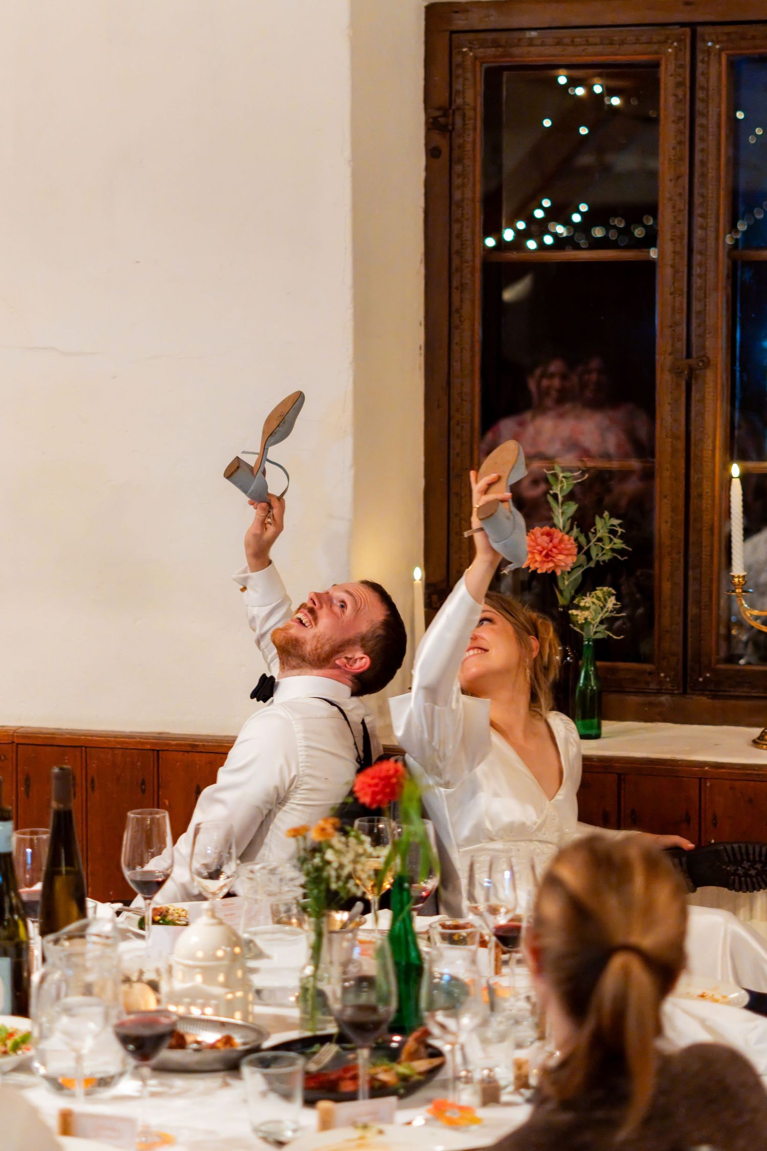 Bride and groom laughing and holding up shoes during a fun wedding reception game at a candlelit dinner table.
