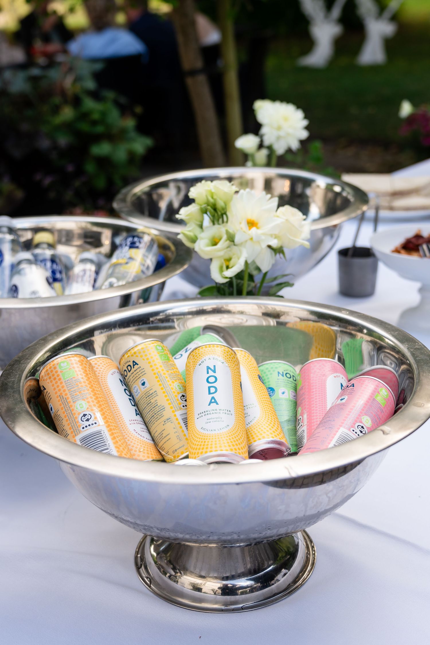 Colorful NODA soda cans chilling in a silver bowl at an outdoor party table with white flowers.