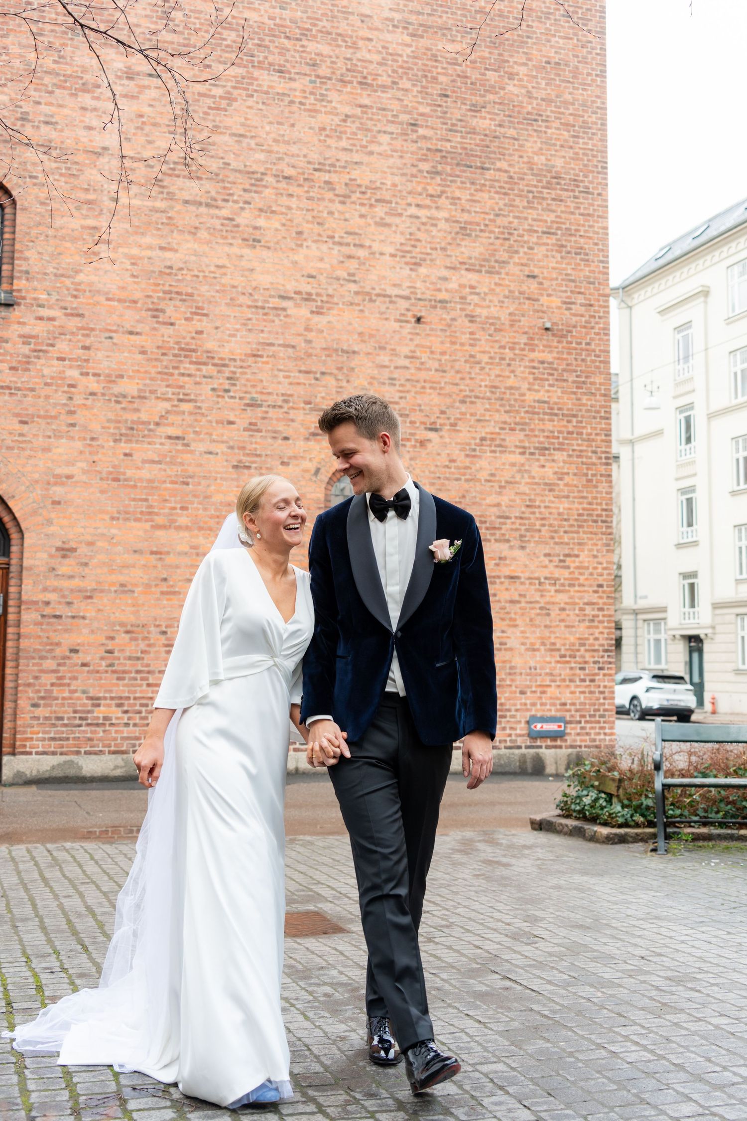A smiling bride in white gown and groom in navy tuxedo walk hand in hand on cobblestone street near brick building.