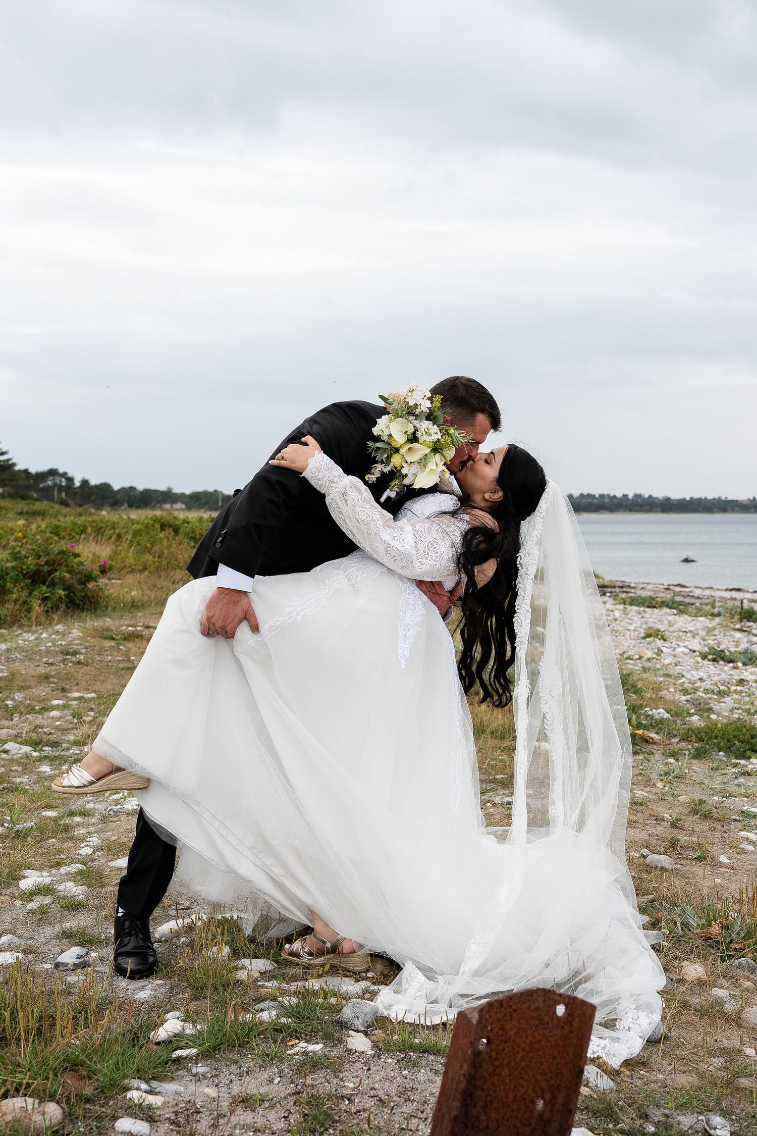 Groom dips bride for a romantic kiss near a scenic waterfront, bride holding a white bouquet in a flowing gown with veil.