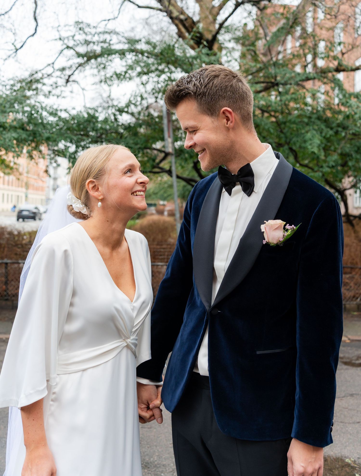 A bride in a white dress and groom in a navy velvet tuxedo hold hands and smile at each other outdoors.