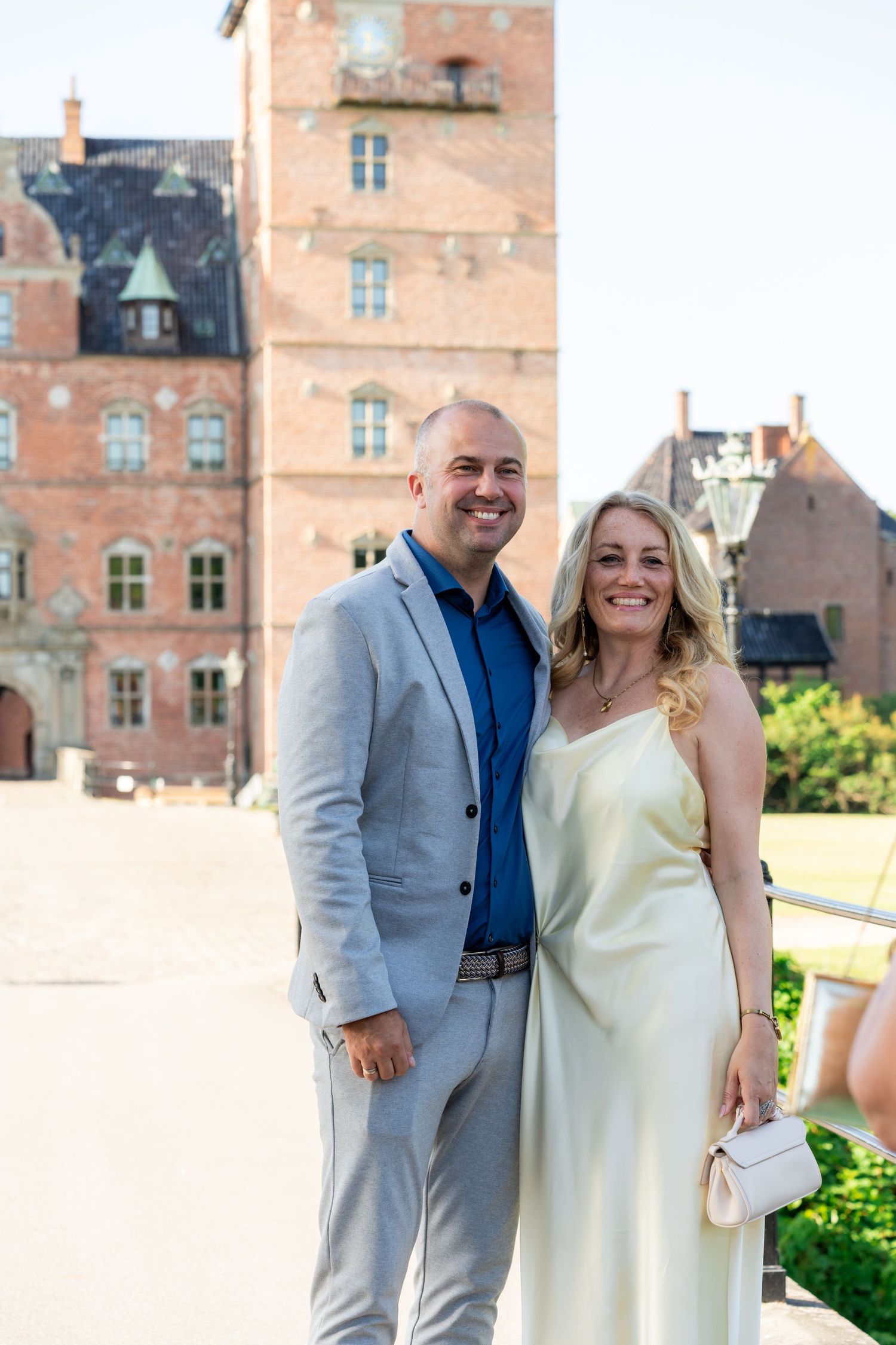 Smiling couple posing in front of a historic red brick castle on a sunny day, man in gray suit, woman in yellow dress.