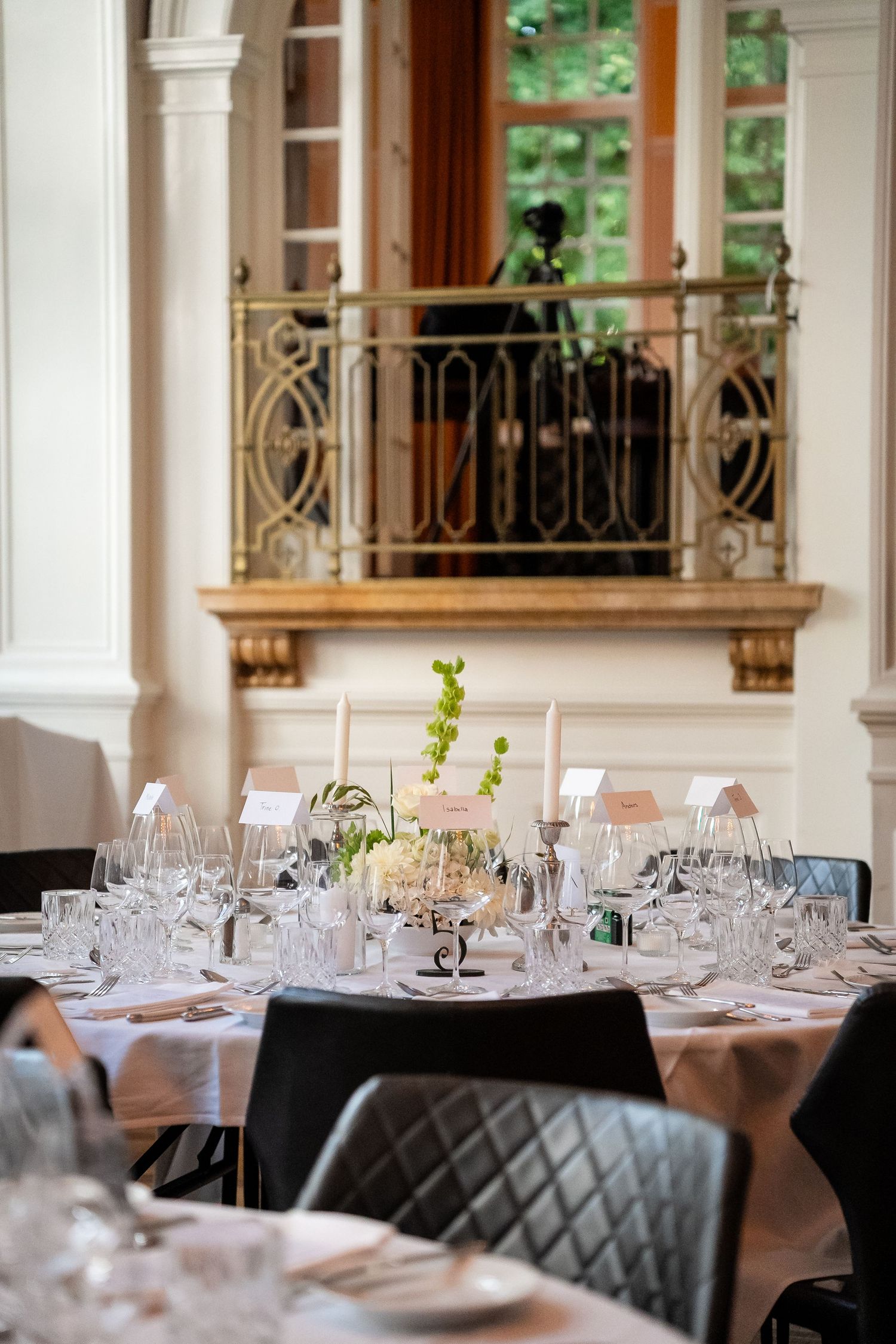 Elegant banquet hall with round tables set with crystal glasses, white linens, floral centerpieces, and ornate gold balcony.