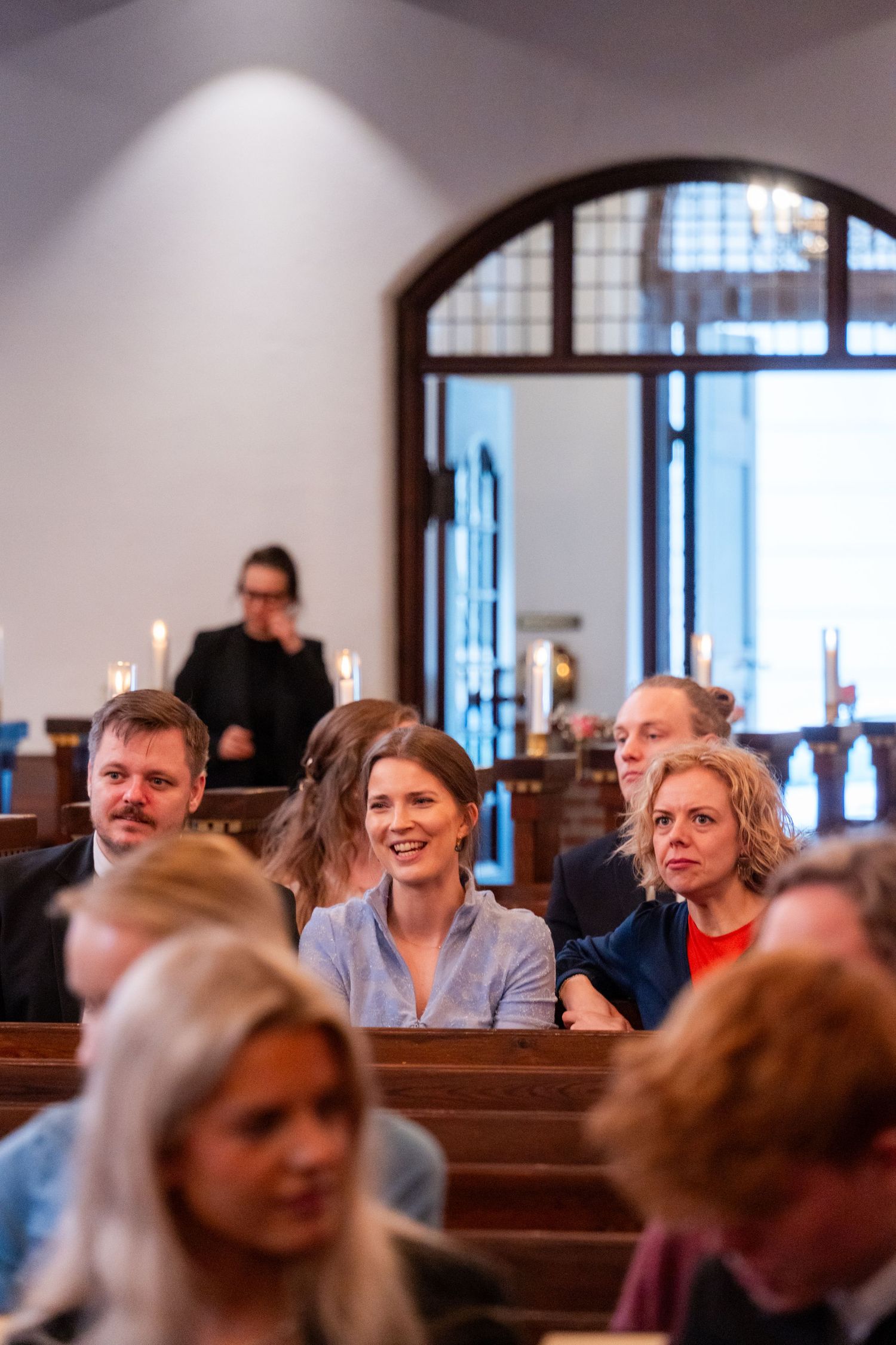 Congregation seated in wooden church pews smiling during a service, with candles and arched windows in background.