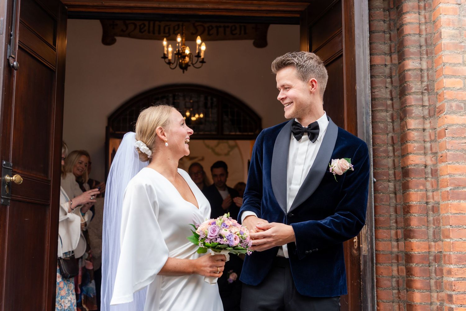 Bride and groom exchanging rings at church entrance, bride in white dress with veil holding bouquet, groom in navy tuxedo.