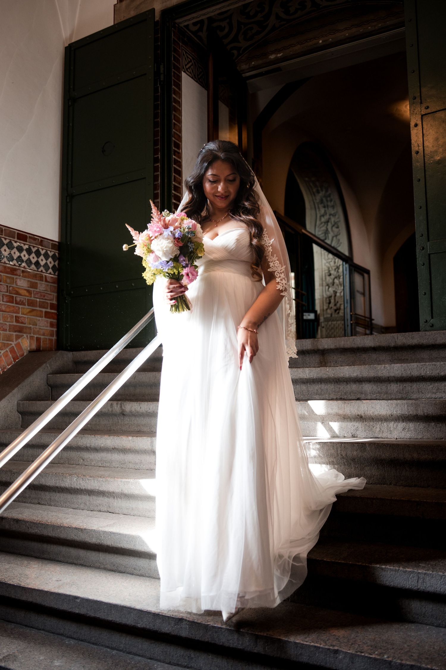 Bride in flowing white gown descending vintage brick stairs with floral bouquet.