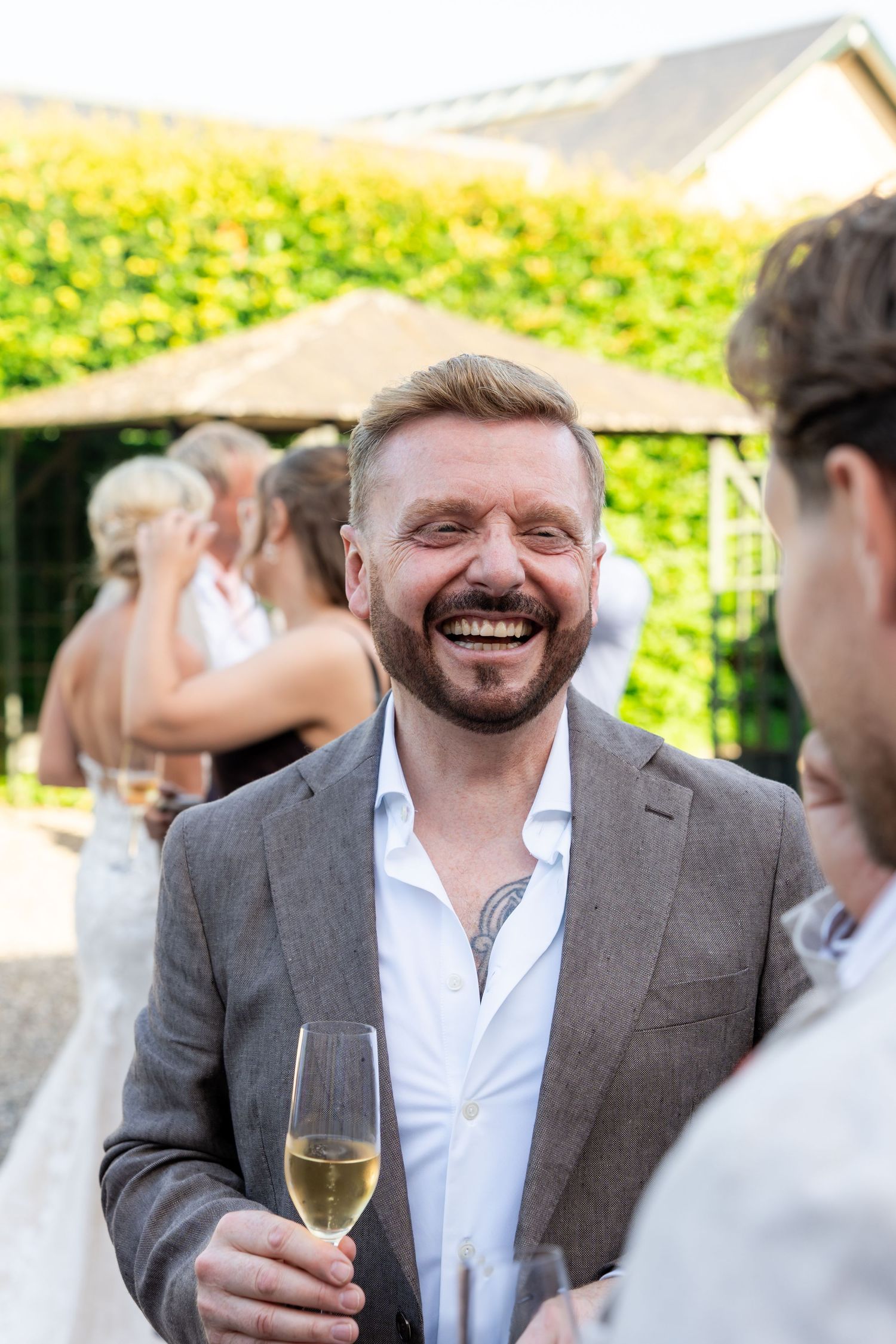 Laughing man in gray blazer holding champagne glass at outdoor wedding reception with guests in background.