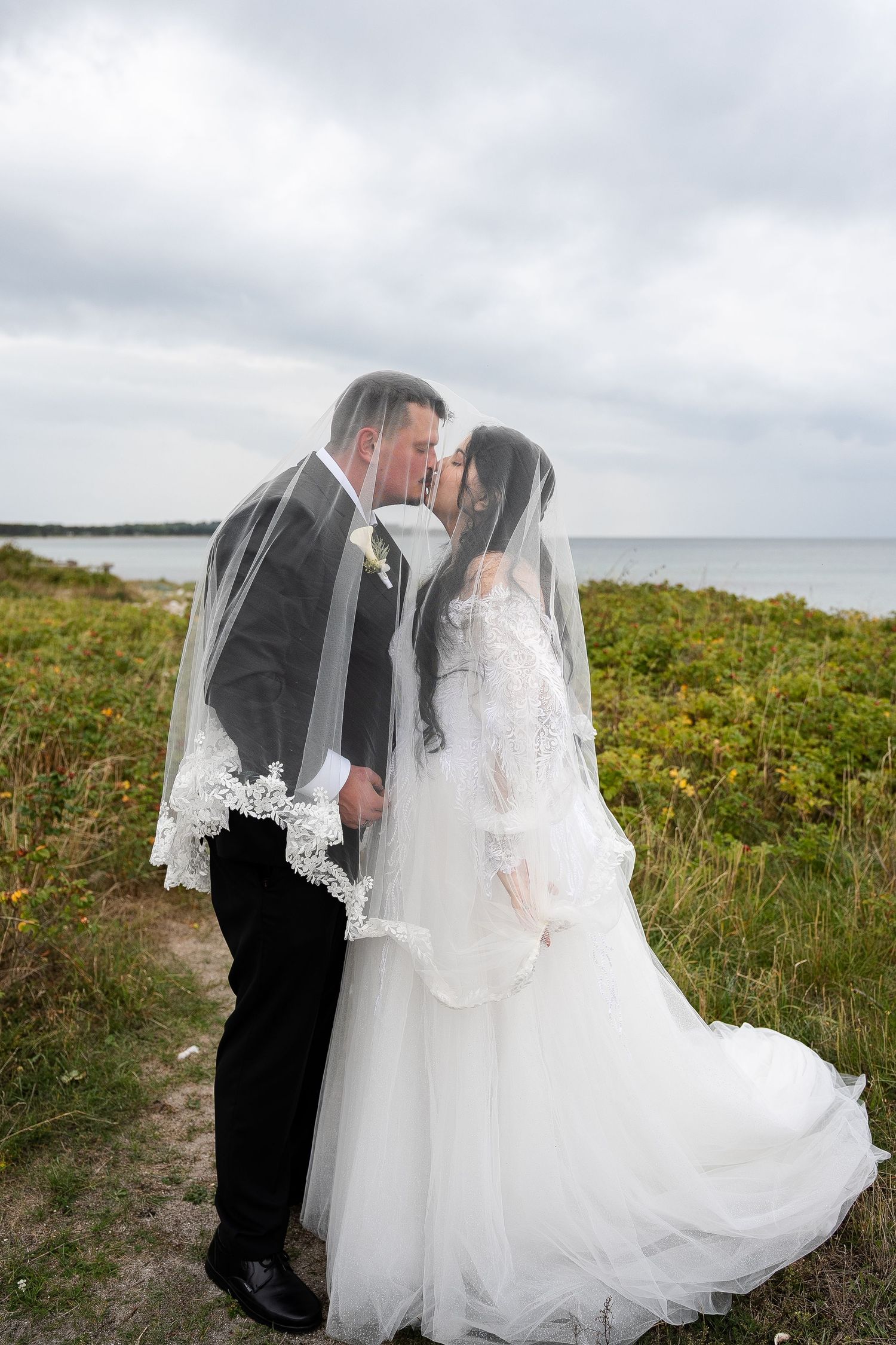 Bride and groom kissing under wedding veil by seaside, outdoor coastal wedding portrait.