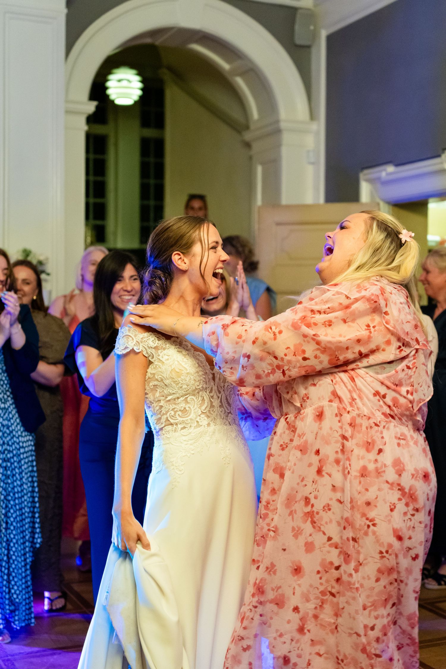 Bride in white lace gown laughing joyfully with guest in floral dress during wedding reception dance floor celebration.