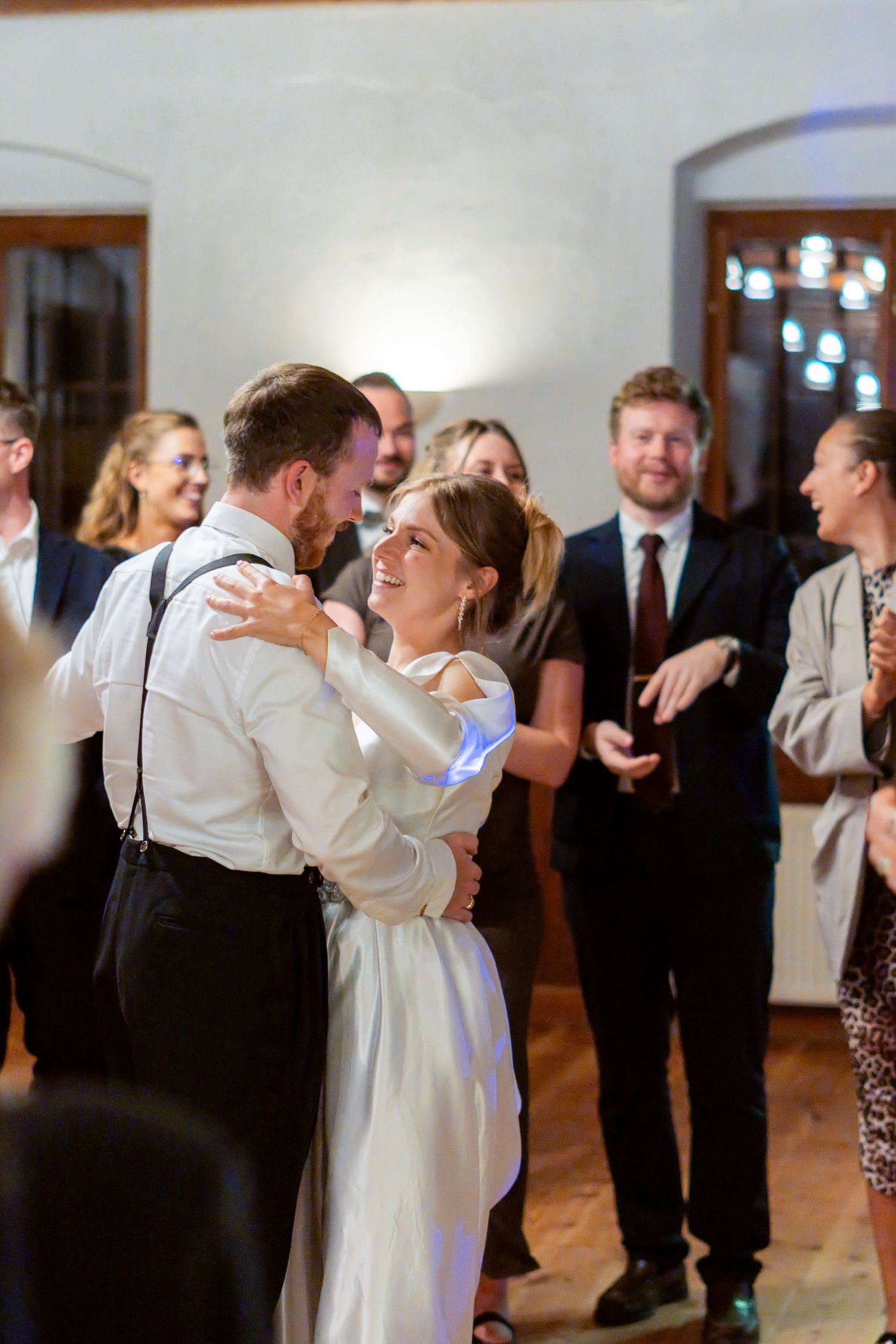 A joyful bride and groom share their first dance surrounded by smiling wedding guests in an elegant venue.