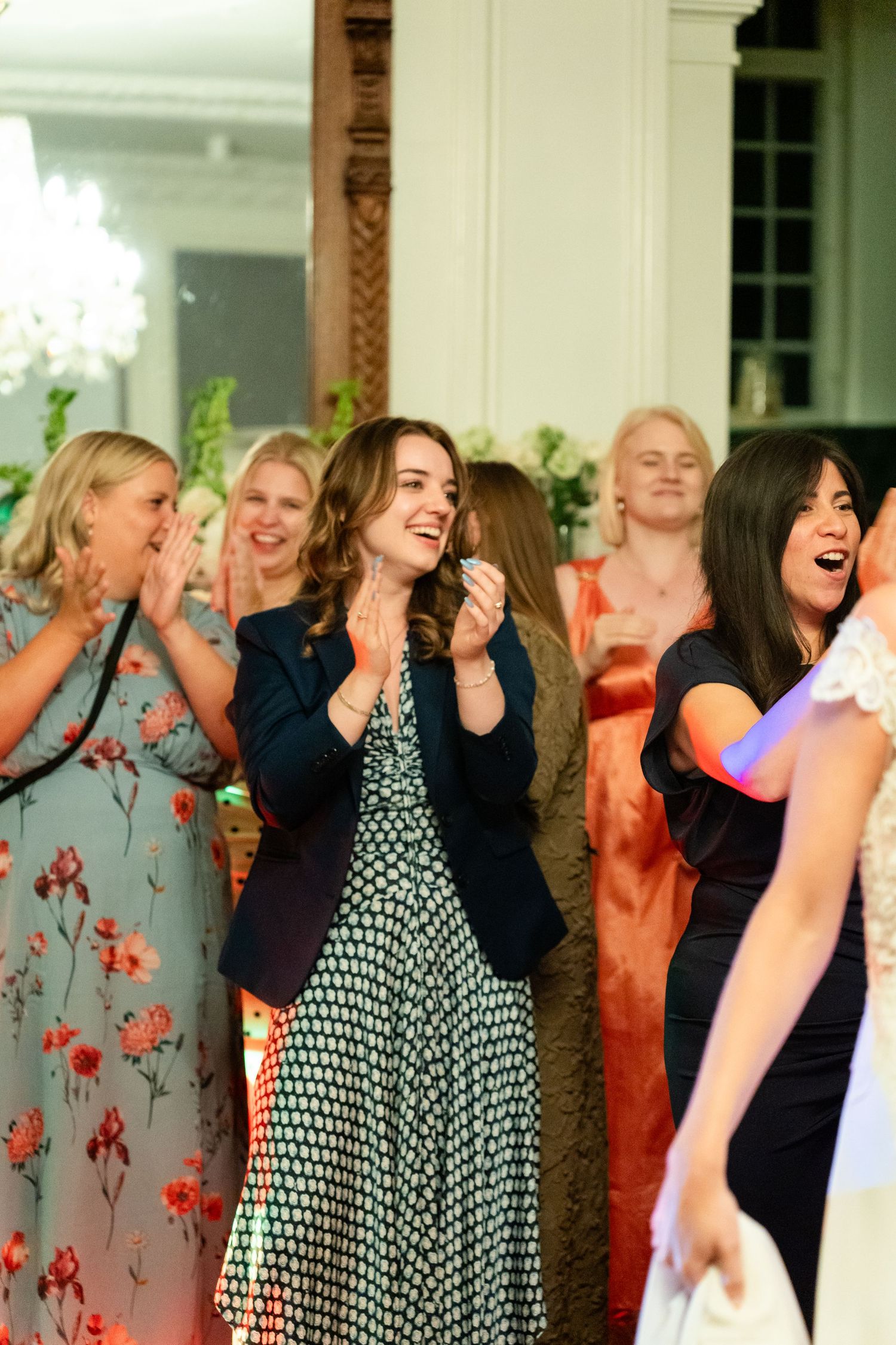 Guests clapping and laughing at a wedding reception in an elegant venue, wearing floral and formal dresses.