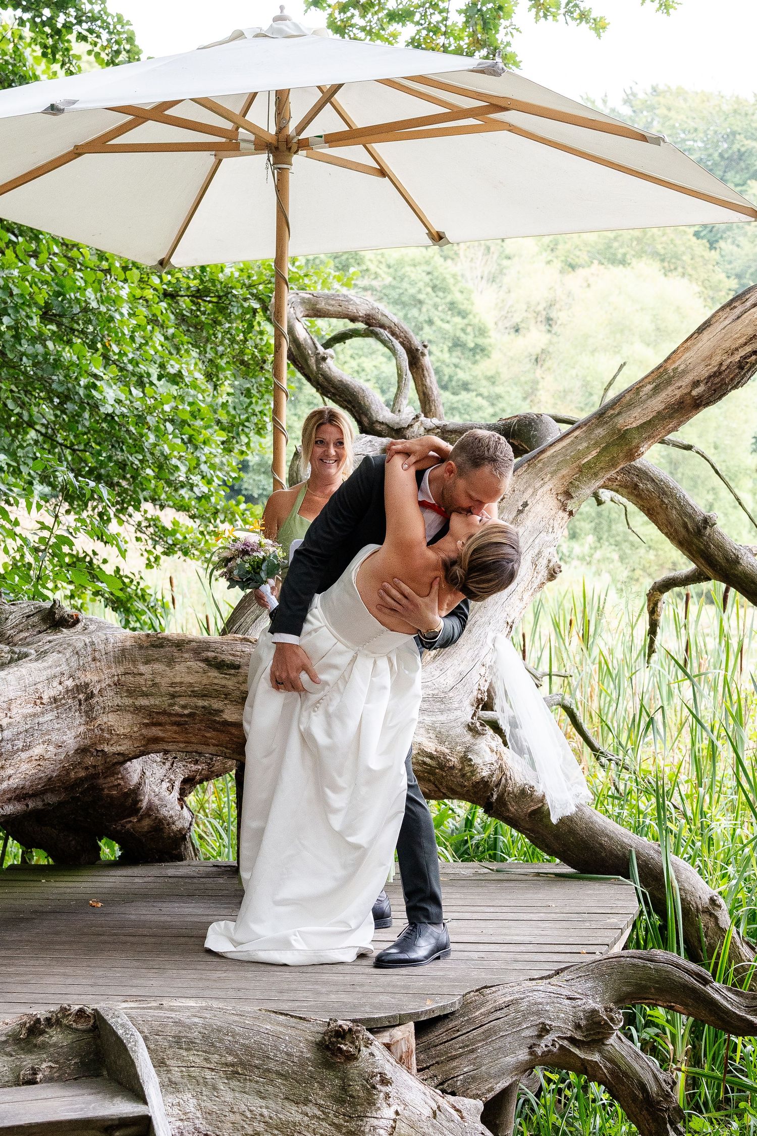 Bride and groom share a romantic dip kiss under a white umbrella on a wooden deck surrounded by lush greenery.