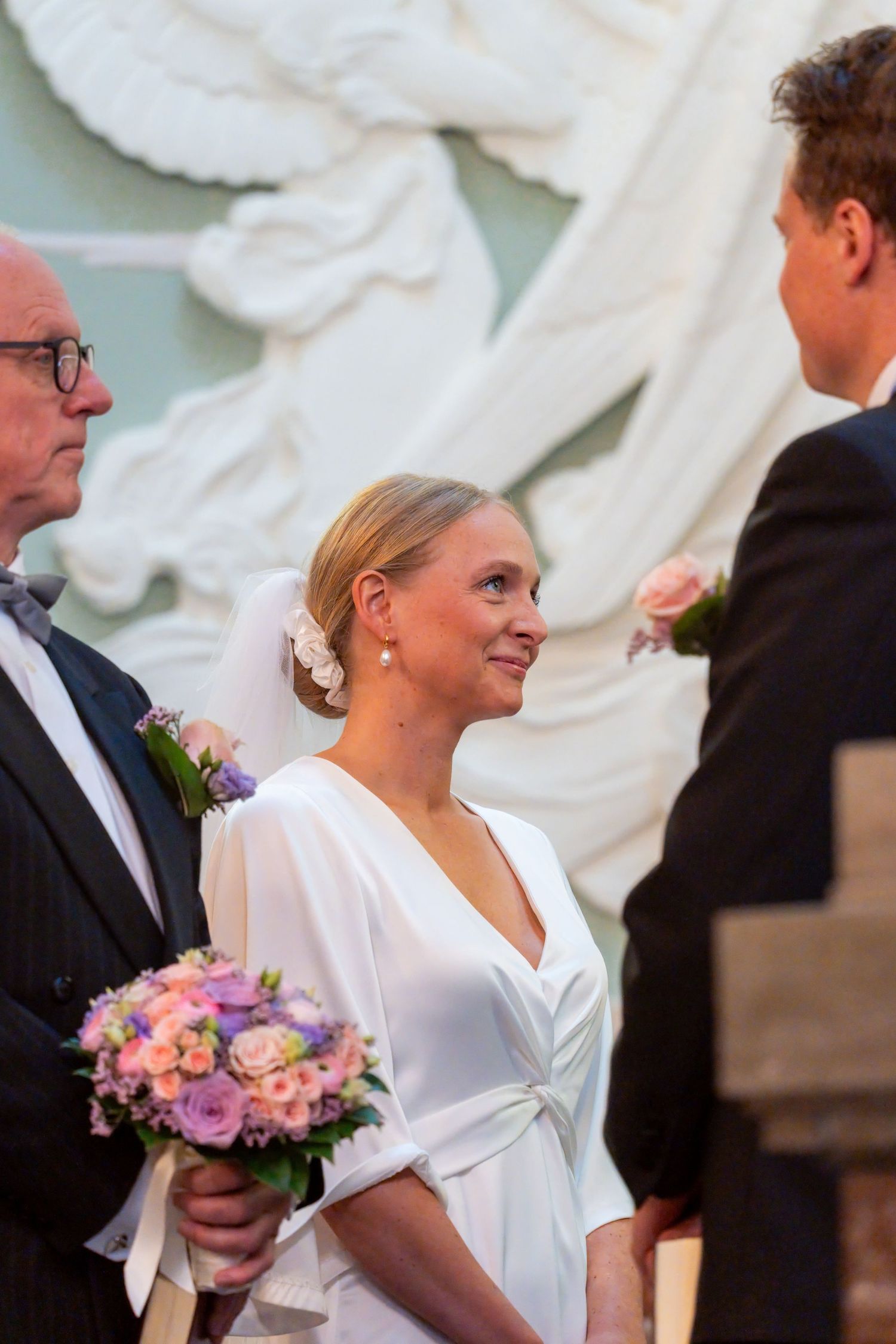 A bride in a white dress stands between two men, one of whom appears to be the groom, at what seems to be a wedding ceremony.