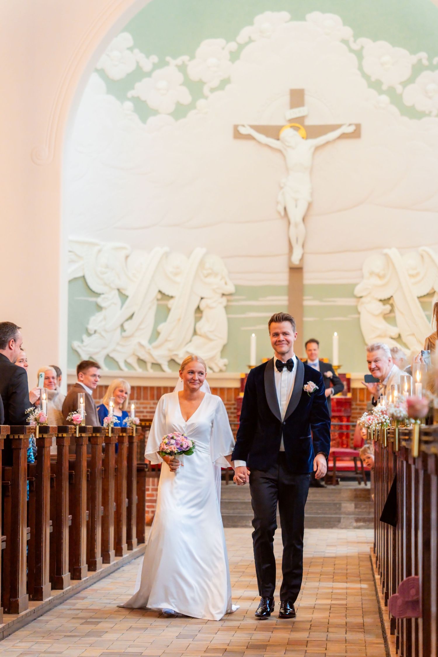 A bride and groom walking down the aisle of a church, with a crucifix visible in the background.