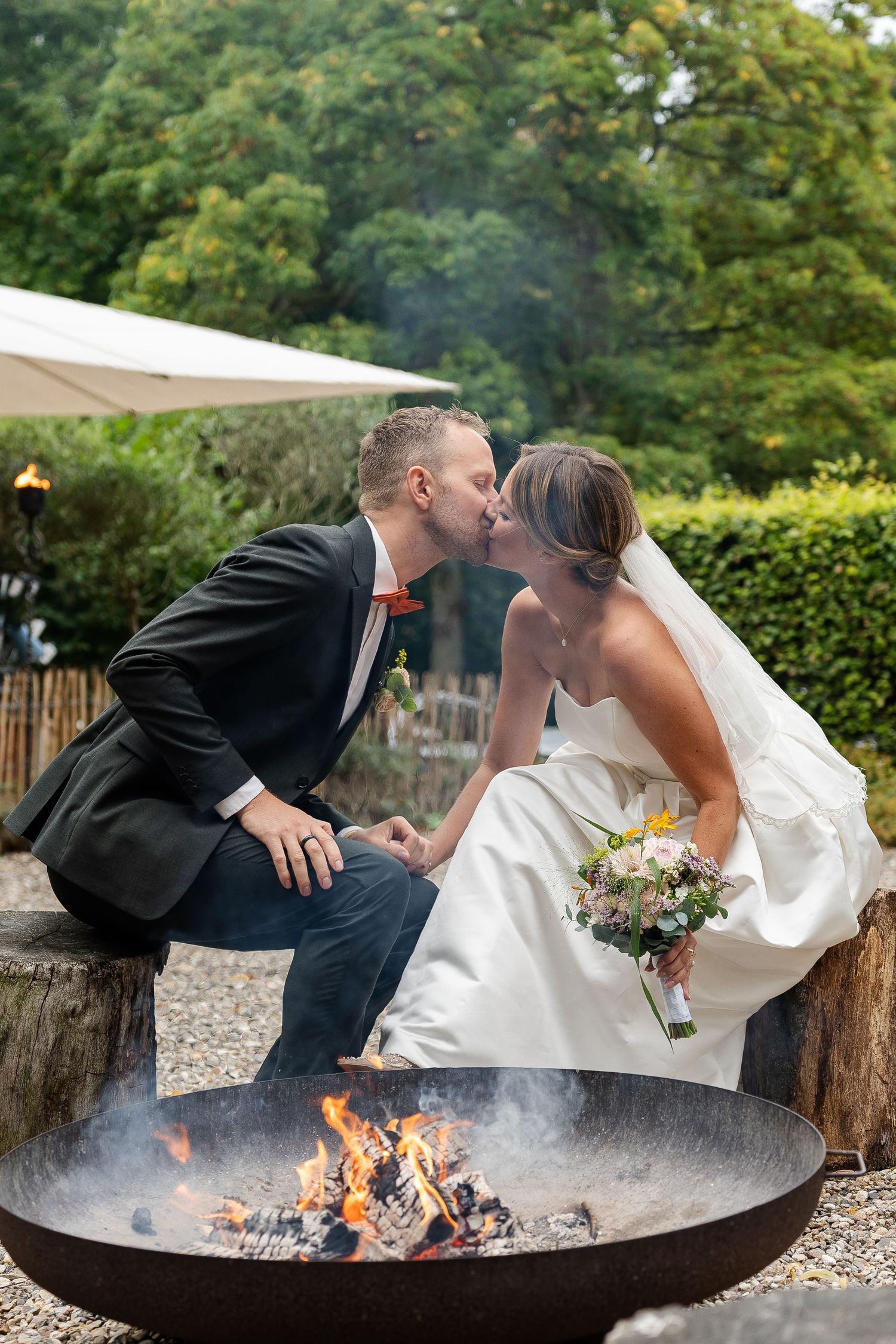 Bride and groom share a romantic kiss while sitting beside a fire pit outdoors on their wedding day.