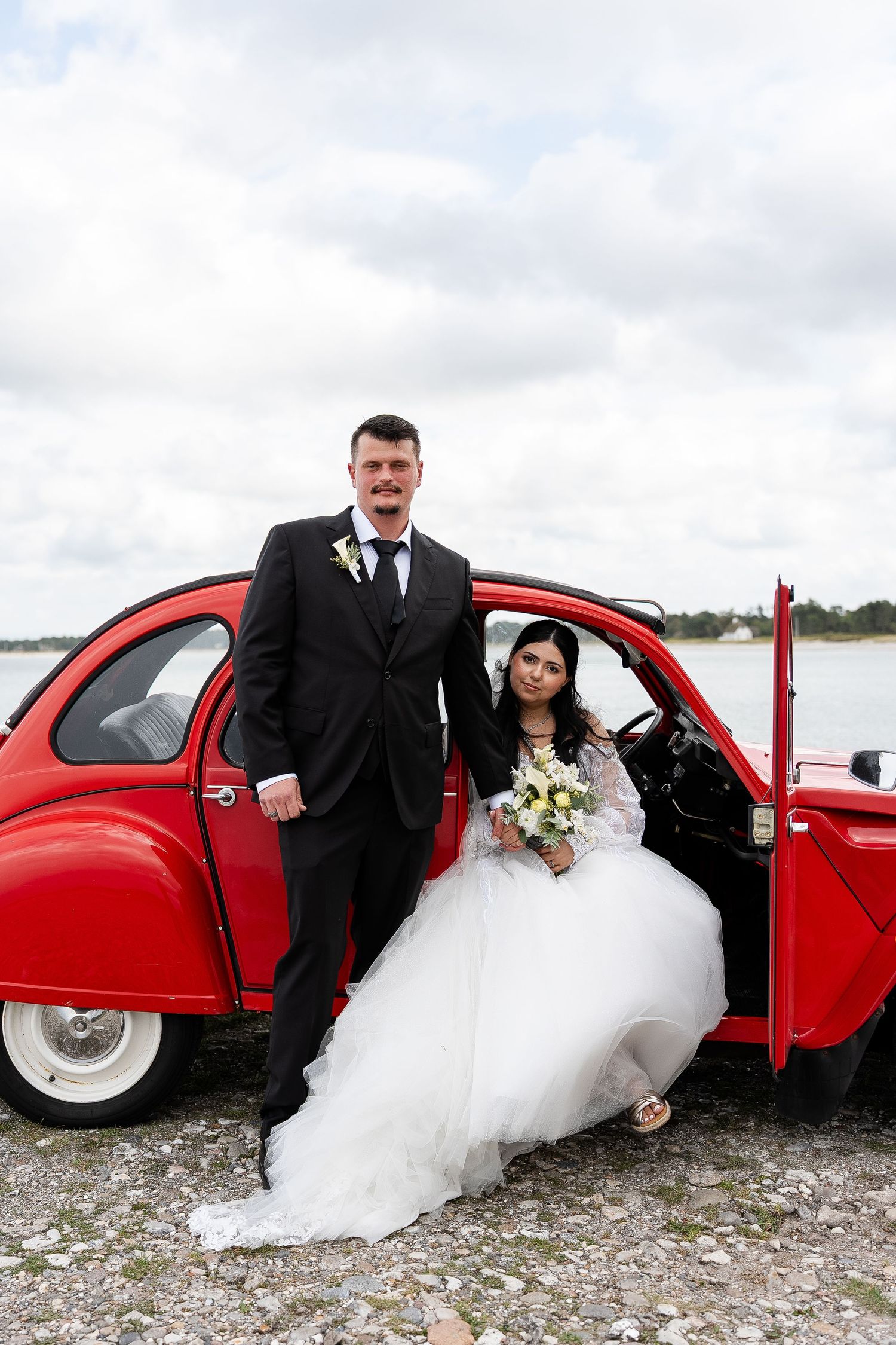 Bride and groom posing beside a classic red Citroen 2CV car on their wedding day, with a waterfront backdrop.