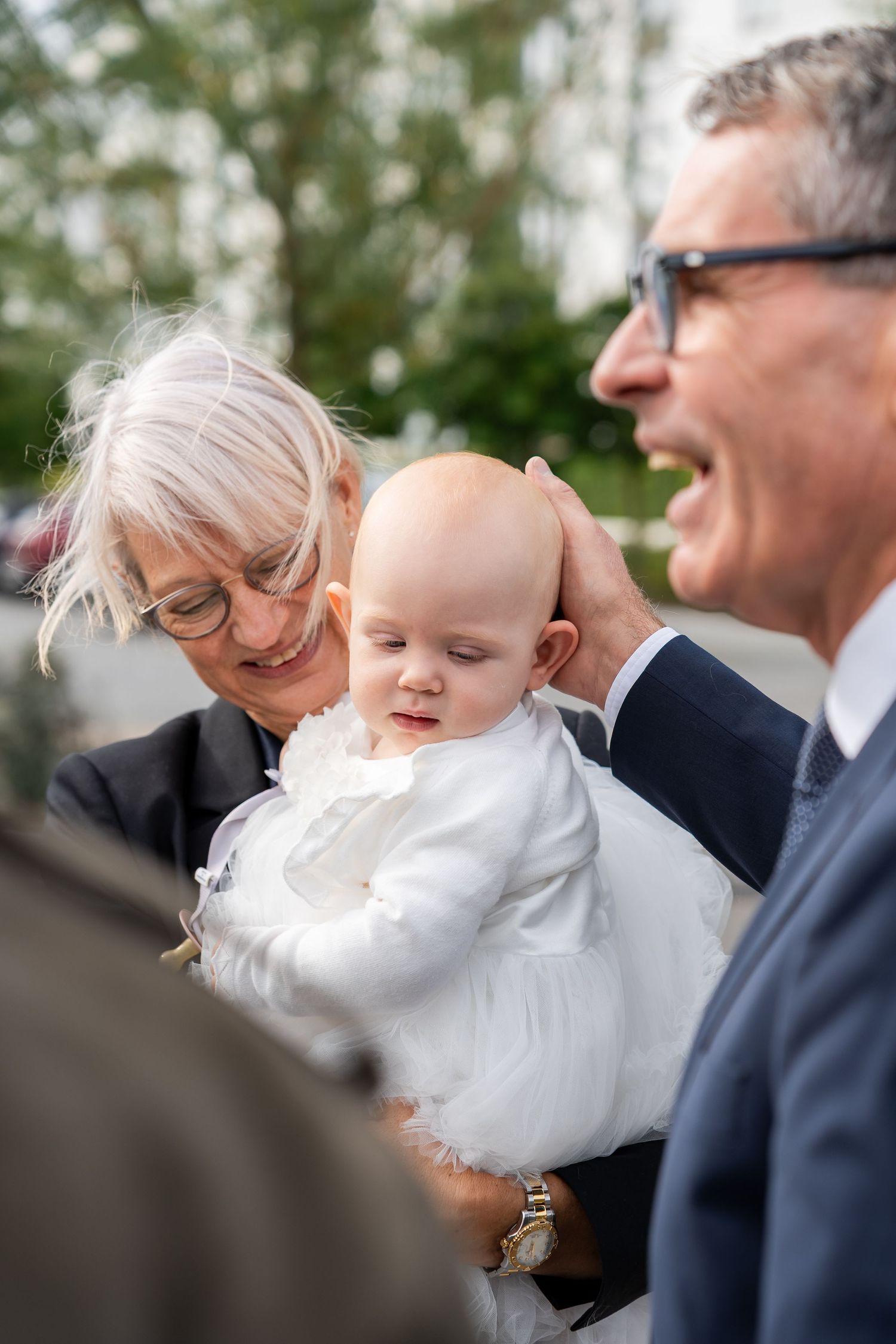 Elderly grandparents lovingly holding a baby dressed in white at what appears to be a christening or baptism ceremony outdoors.