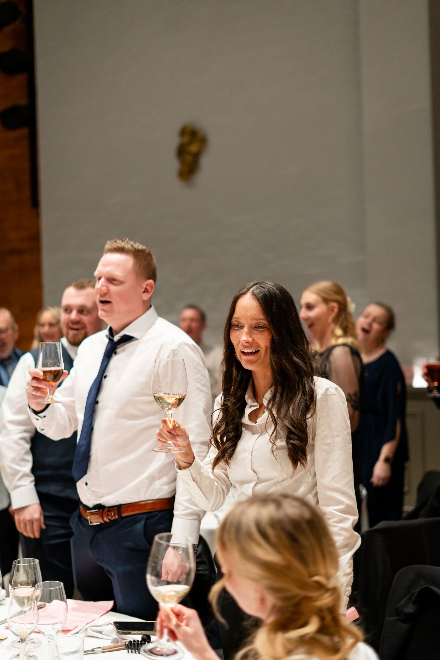 Wedding reception guests laughing and socializing at tables with wine glasses.