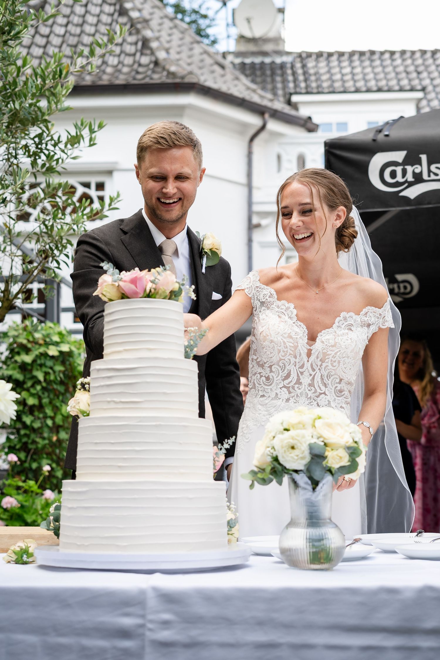 Happy bride and groom laughing while cutting elegant white tiered wedding cake outdoors at a garden reception venue.
