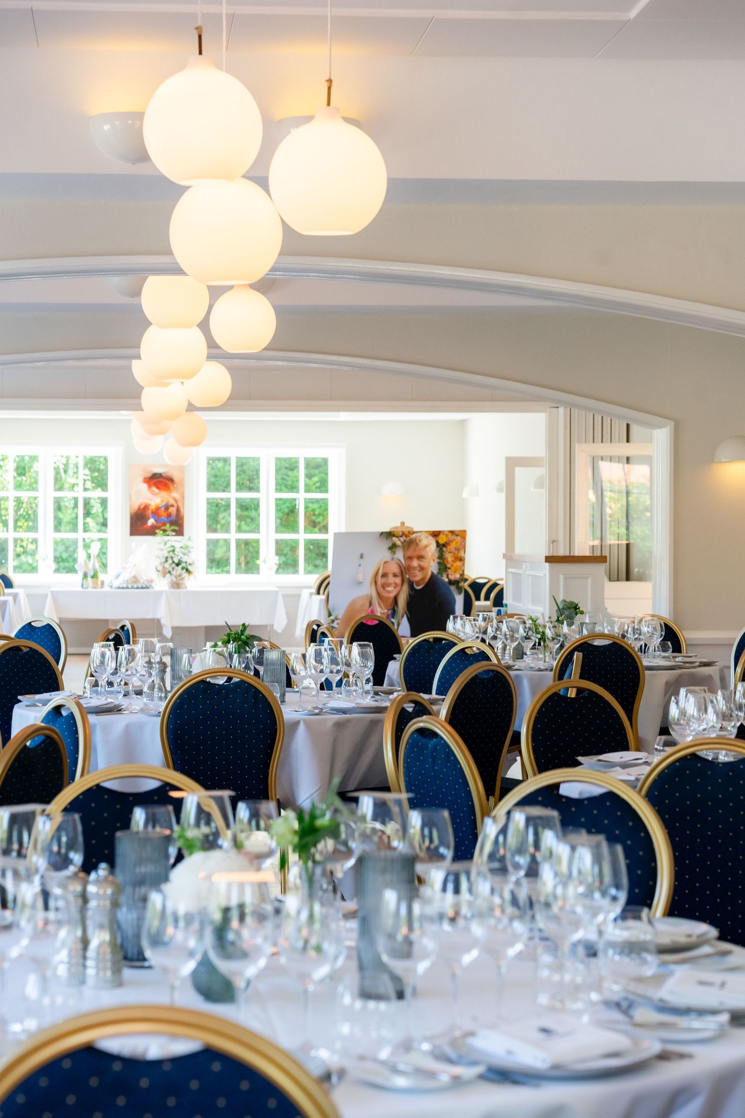 Elegant banquet hall with navy blue chairs, white round pendant lights, and tables set with glassware for a formal event.