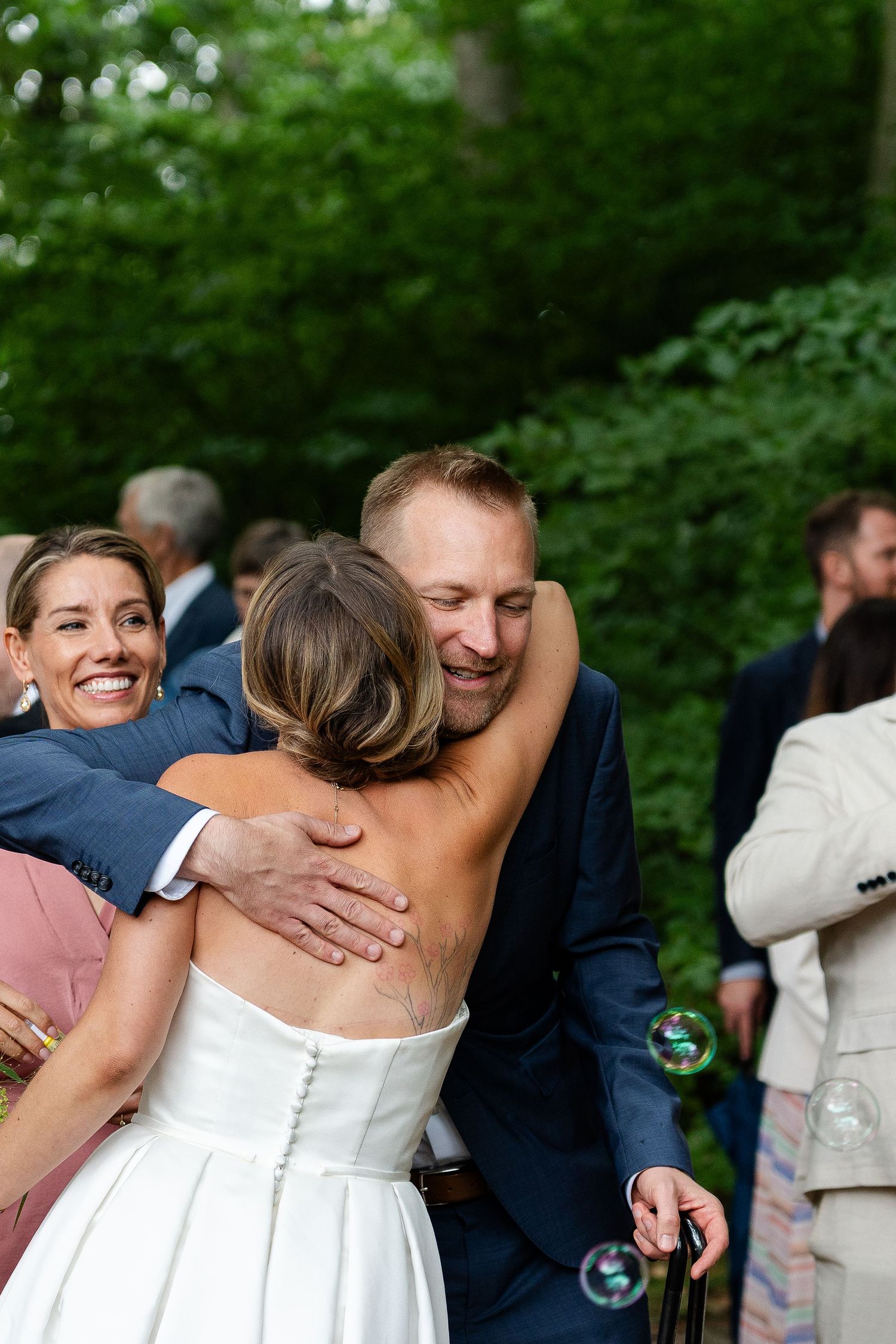 Groom in navy suit hugging bride in backless white gown at outdoor wedding reception surrounded by smiling guests.